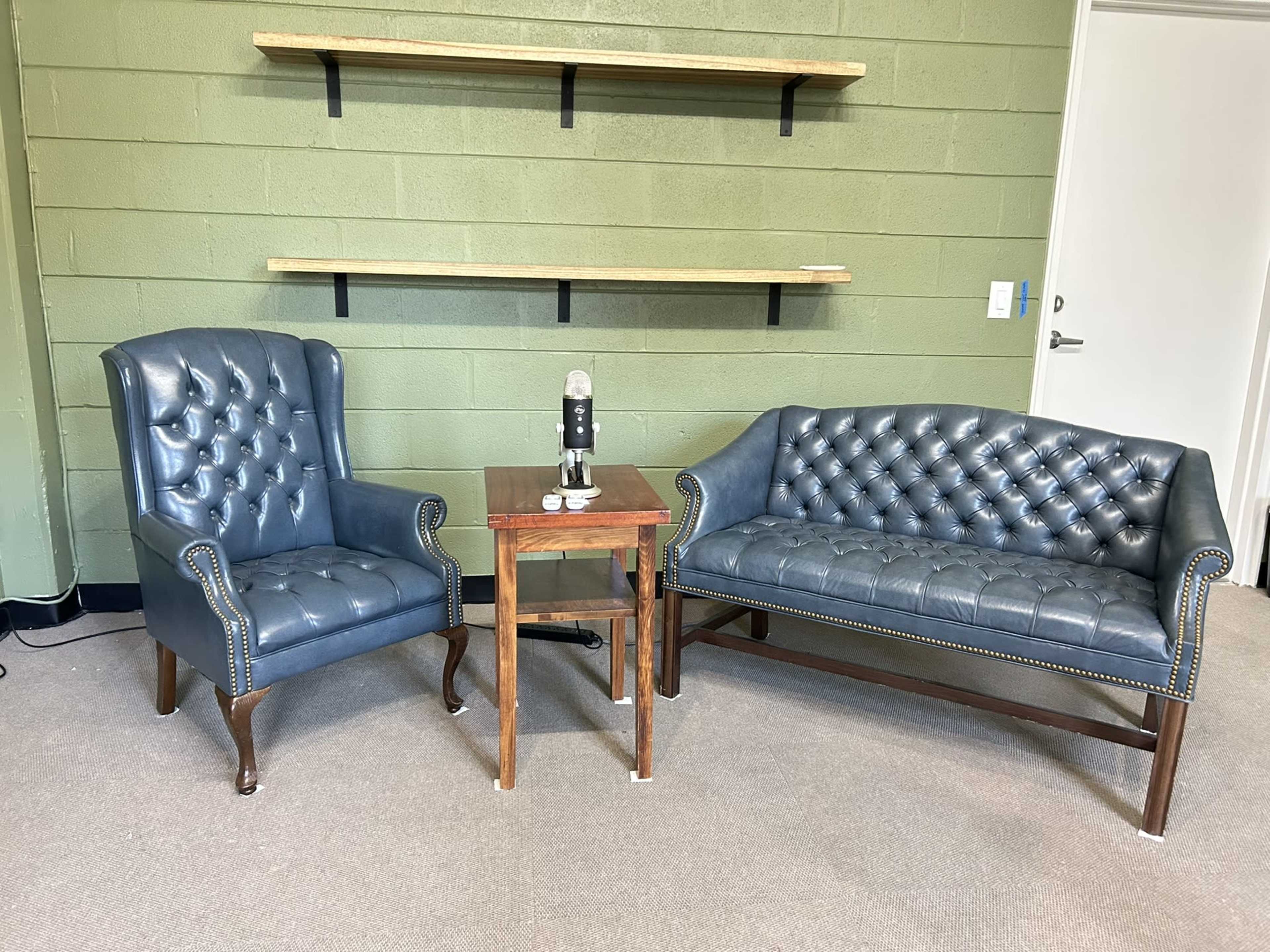 The image shows a room with a green wall featuring two blue leather chairs and a wooden table between them, holding a microphone.