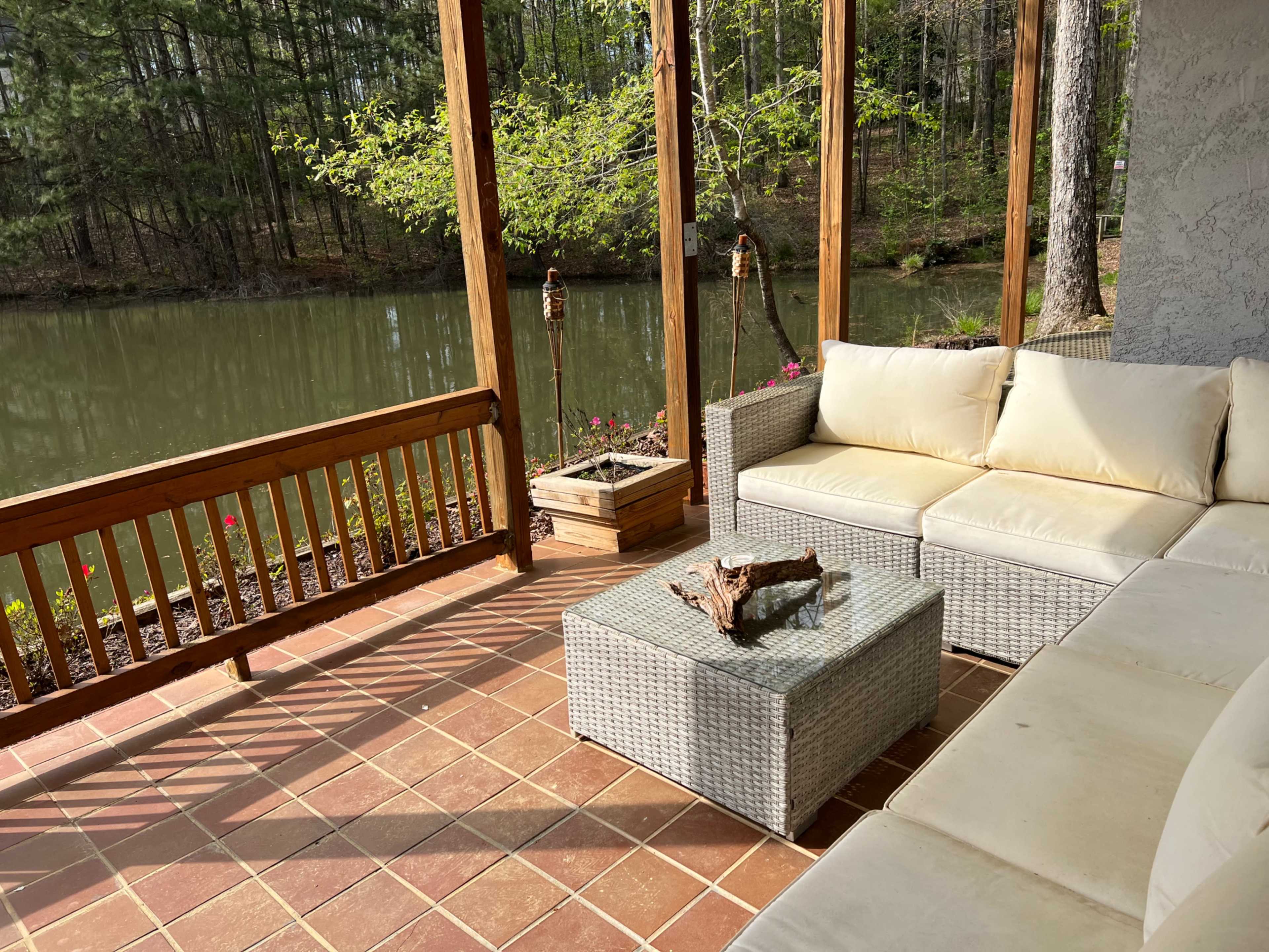 A cozy seating area with a light-colored outdoor sofa and a glass-topped coffee table overlooks a serene pond surrounded by trees.