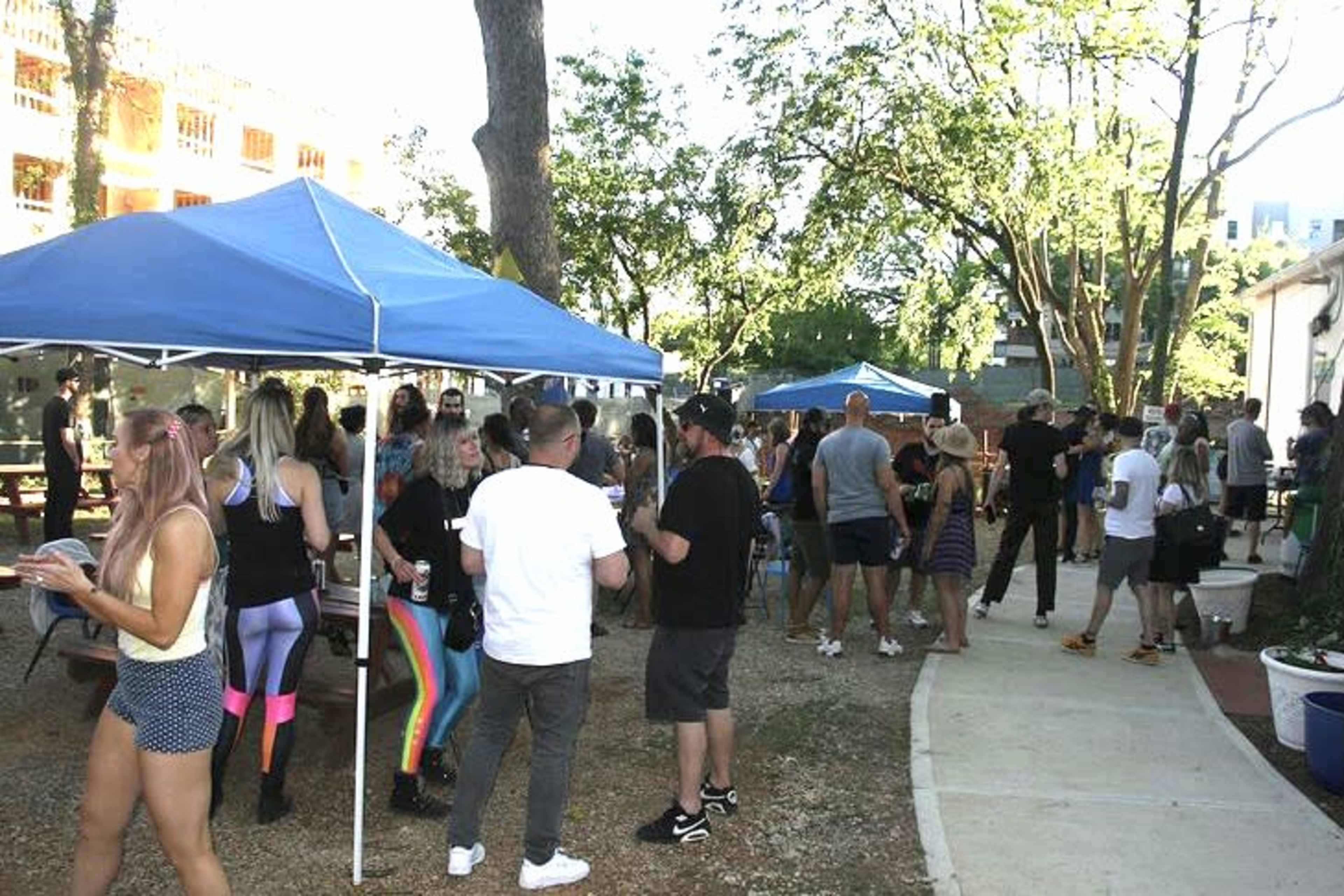 A crowd of people gathers around blue tents in a park setting, with trees and a building in the background.