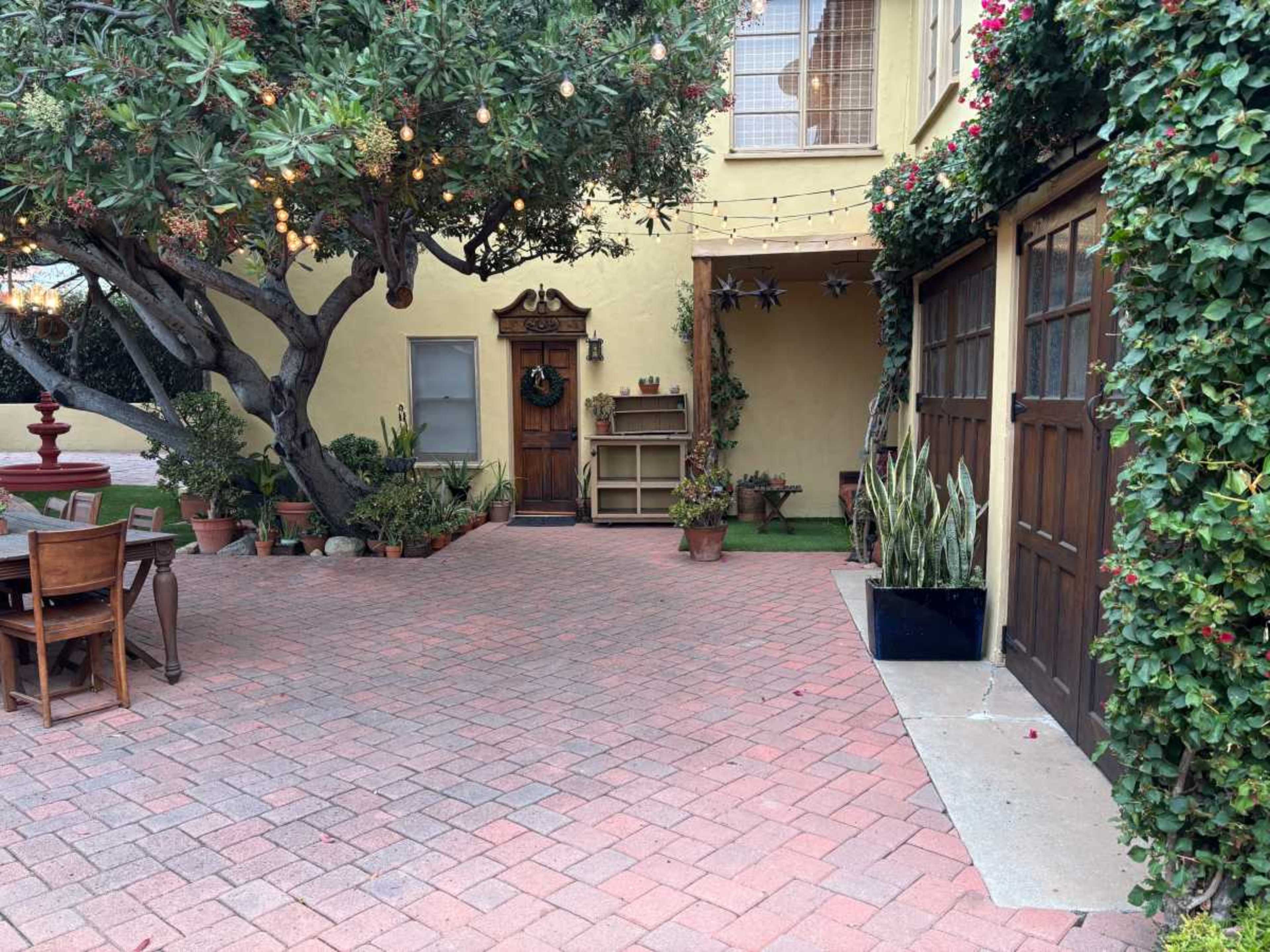 The image shows a brick-paved courtyard with potted plants, a wooden door, and string lights hanging overhead.