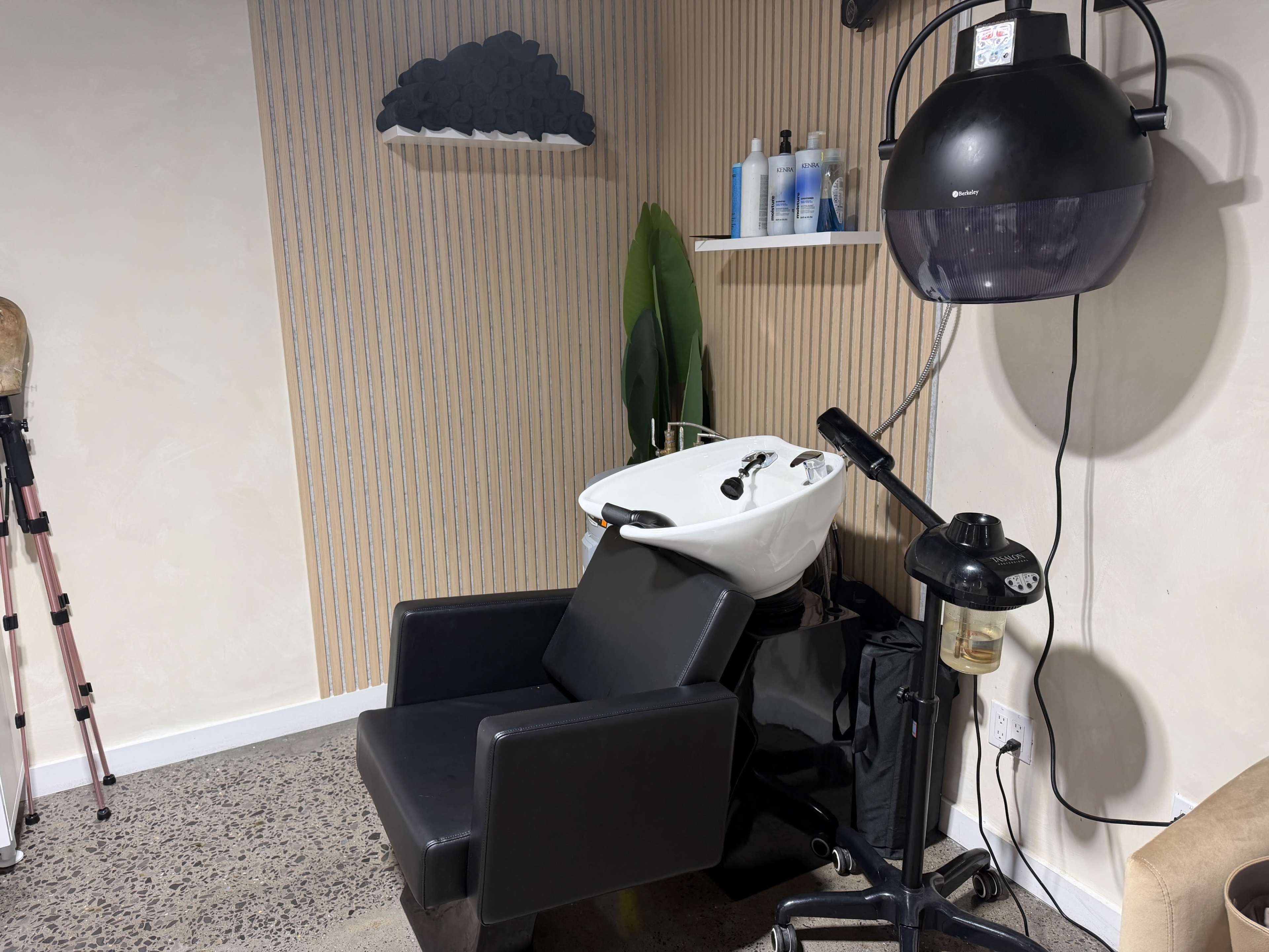 The image shows a salon shampoo station with a white basin, a black salon chair, and a hooded dryer nearby.