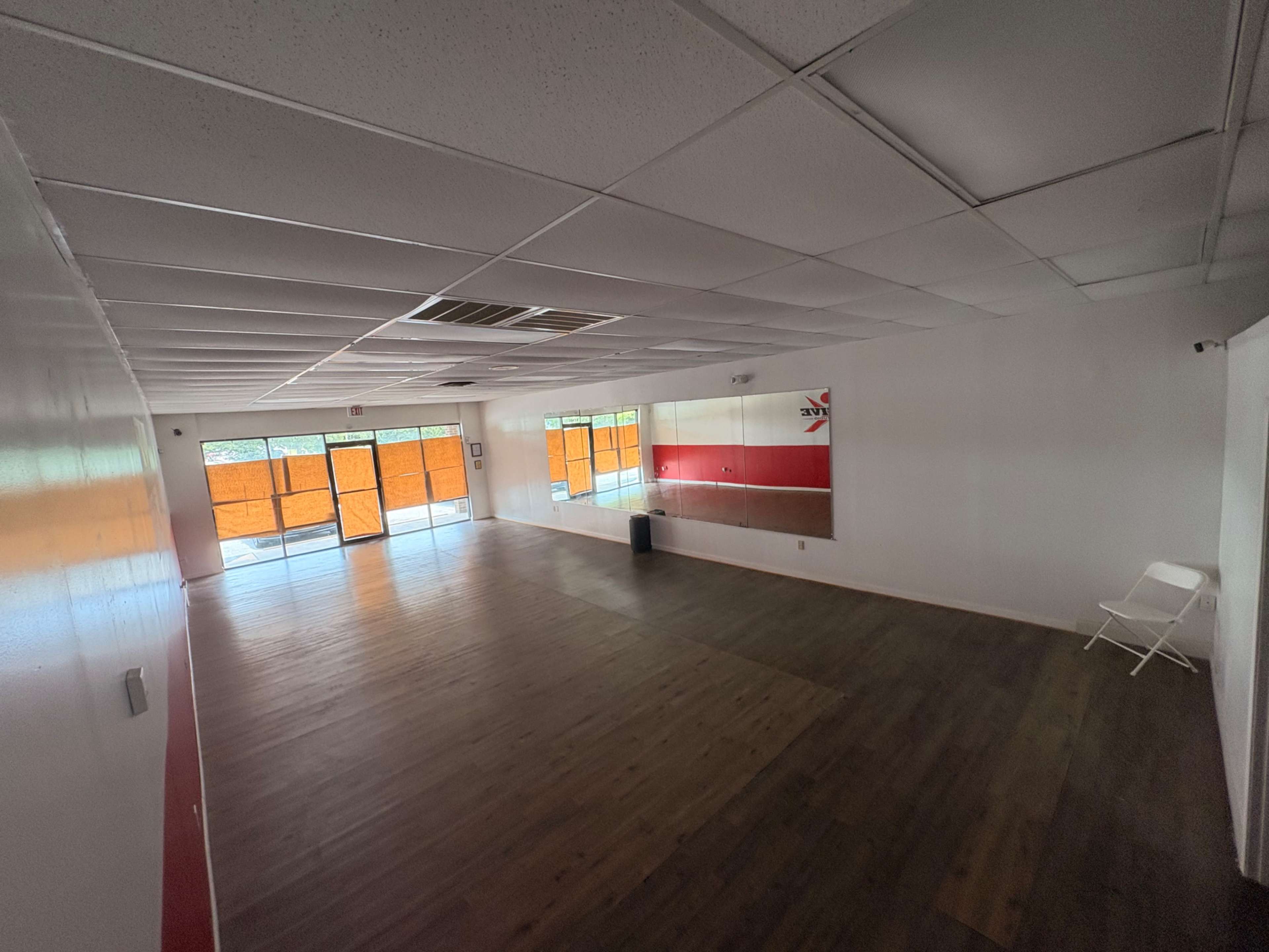 The image shows an empty, brightly lit room with wooden flooring and a large mirror on one wall.