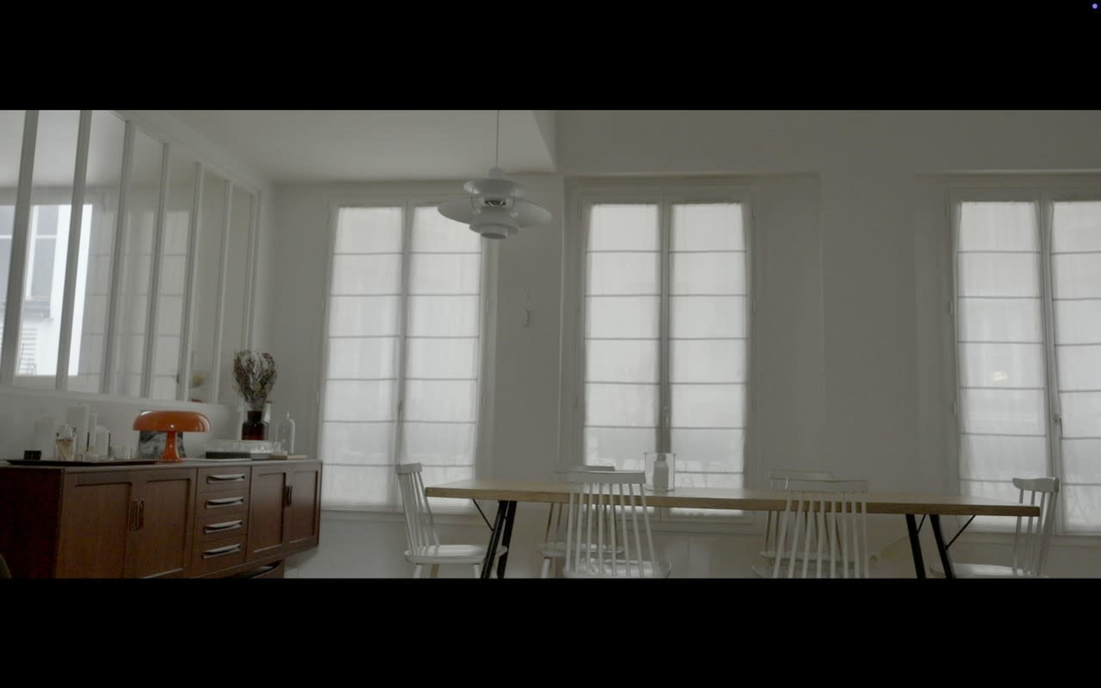 The image depicts a minimalist dining area with a long wooden table, white chairs, and large windows covered by sheer curtains.