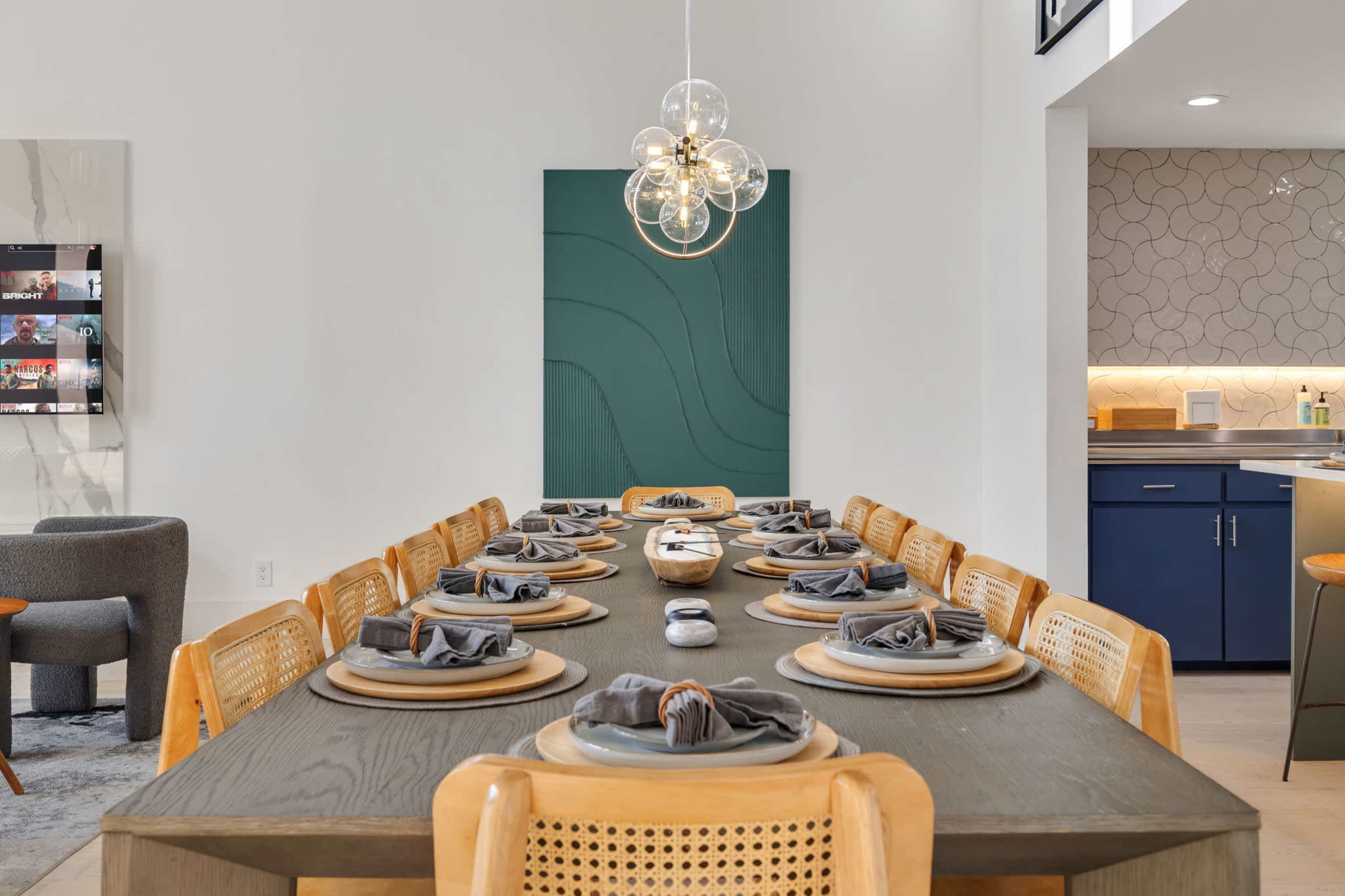 A long dining table is set with plates and napkins, illuminated by a modern pendant light, with a kitchen visible in the background.