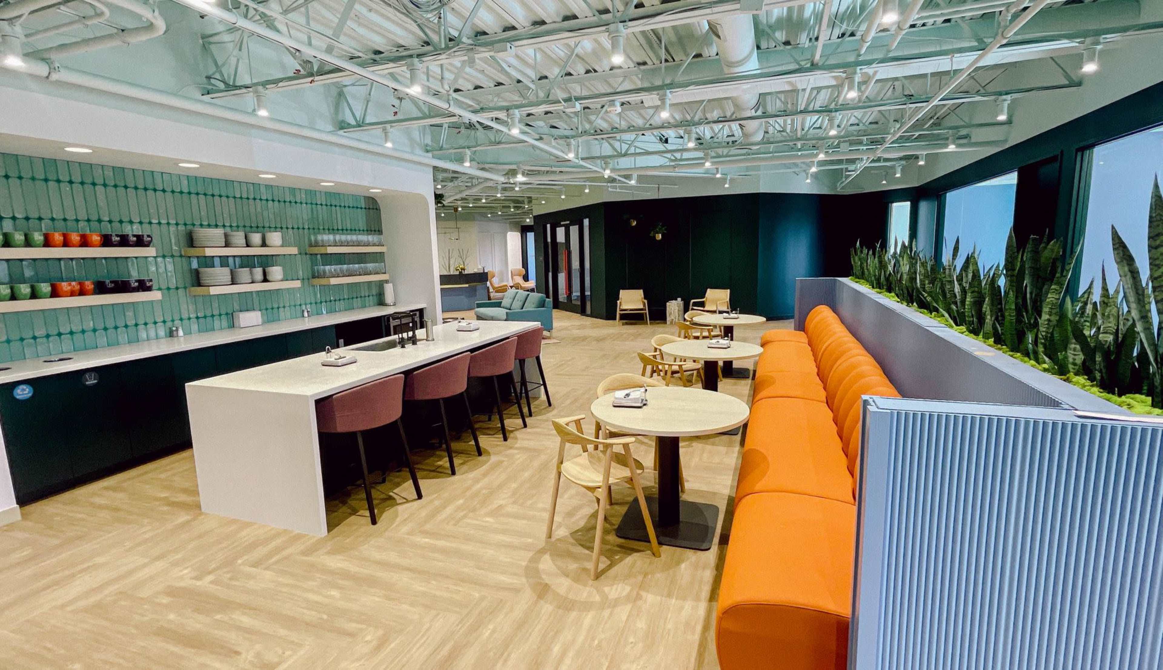 A modern café with a long white counter, orange seating, and several tables arranged around a spacious, well-lit interior featuring plants.