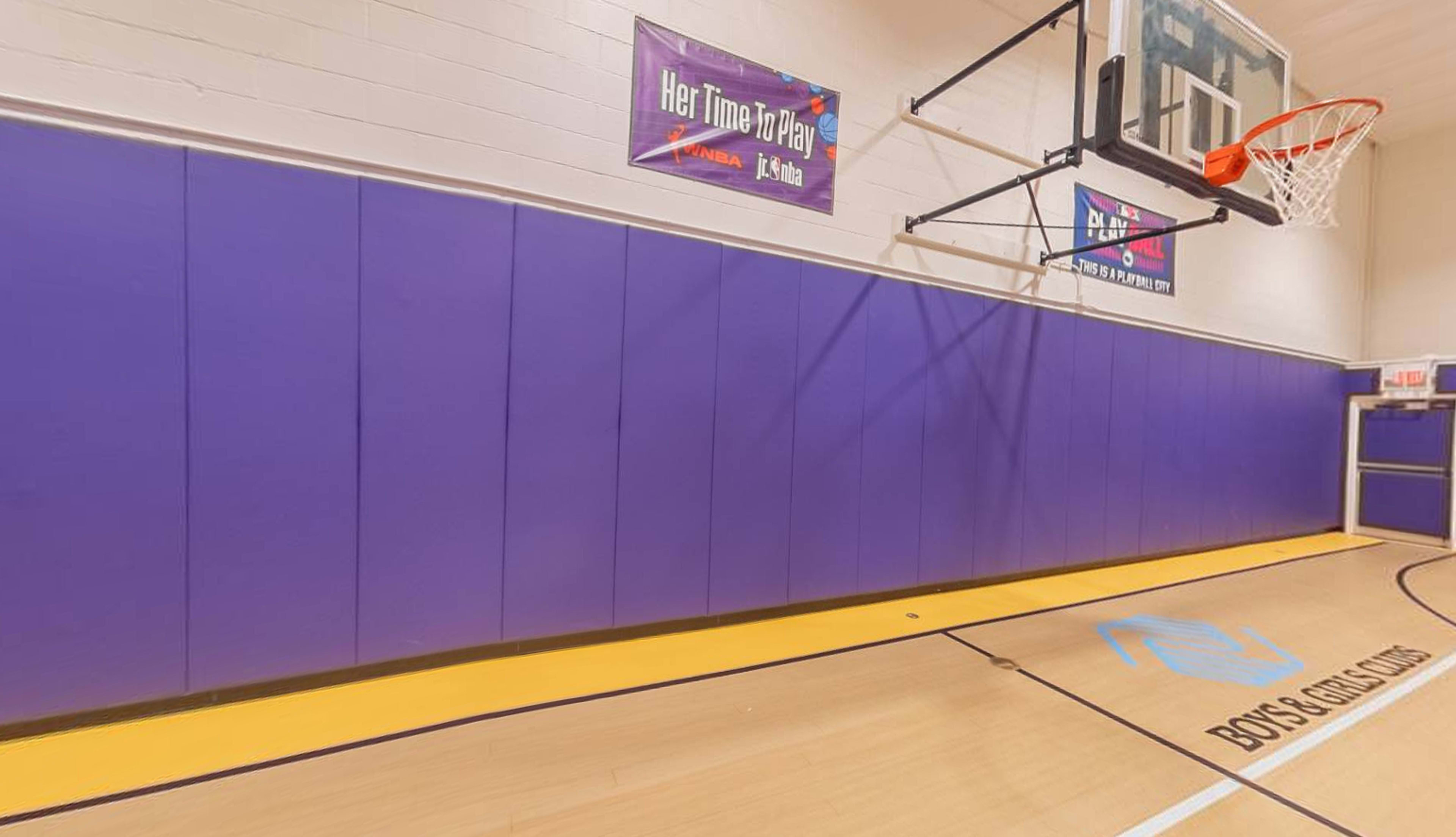 A portion of a gymnasium with purple wall padding, a basketball hoop, and a marked area on the floor.
