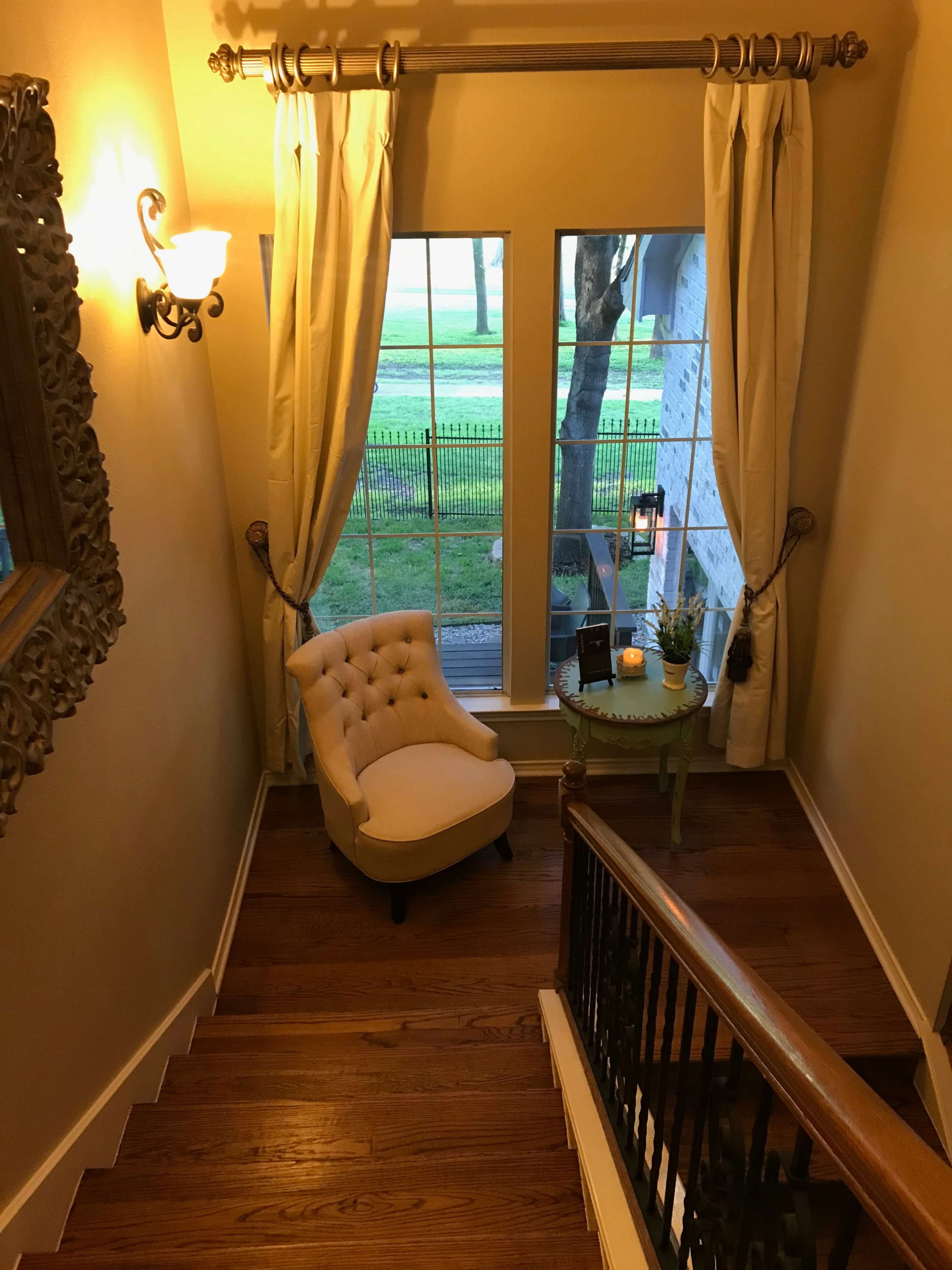 A staircase leads down to a small chair beside a side table, with large windows overlooking a green area outside.
