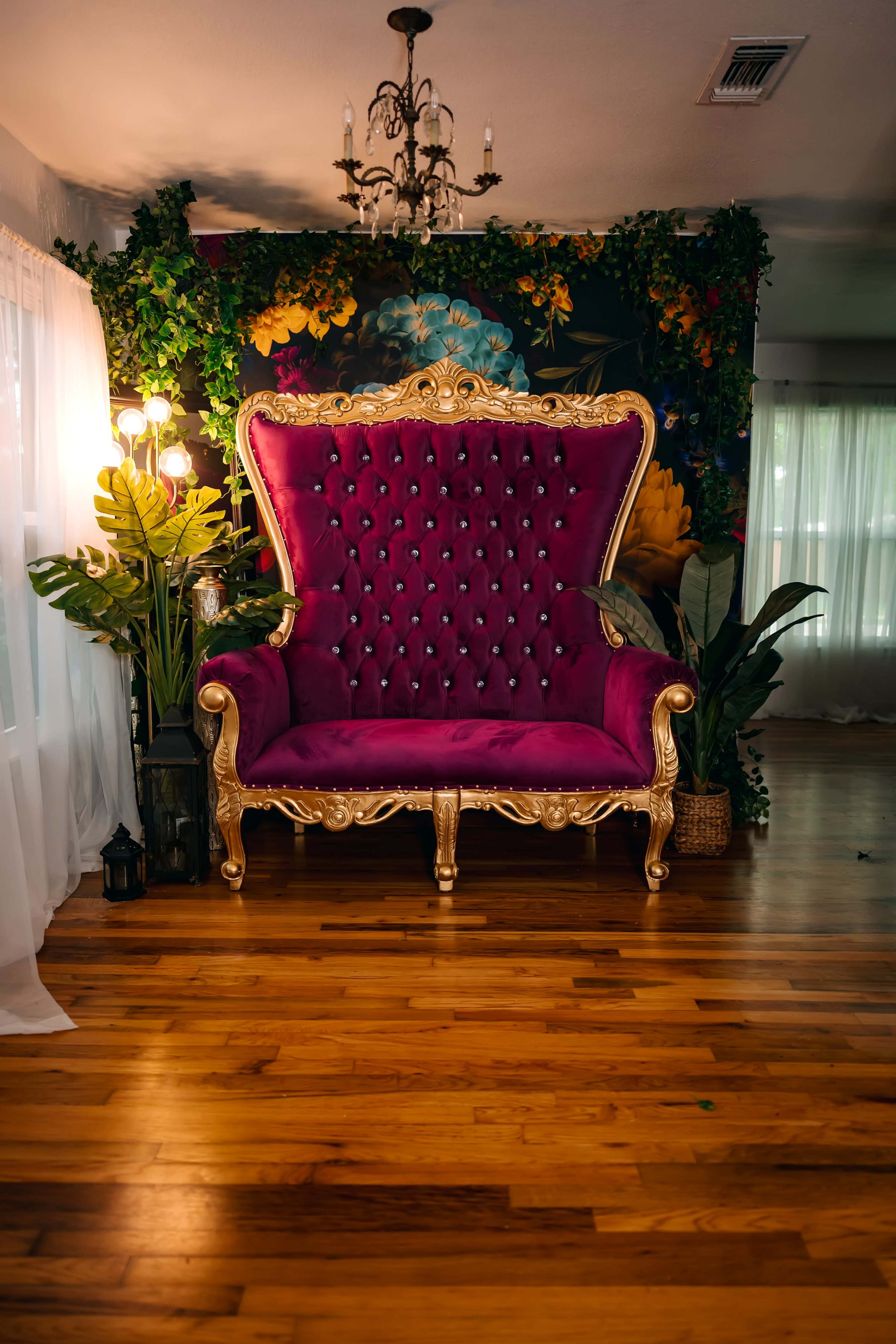 The image features a large, ornate purple chair with a gold frame, positioned against a floral backdrop and surrounded by decorative plants.