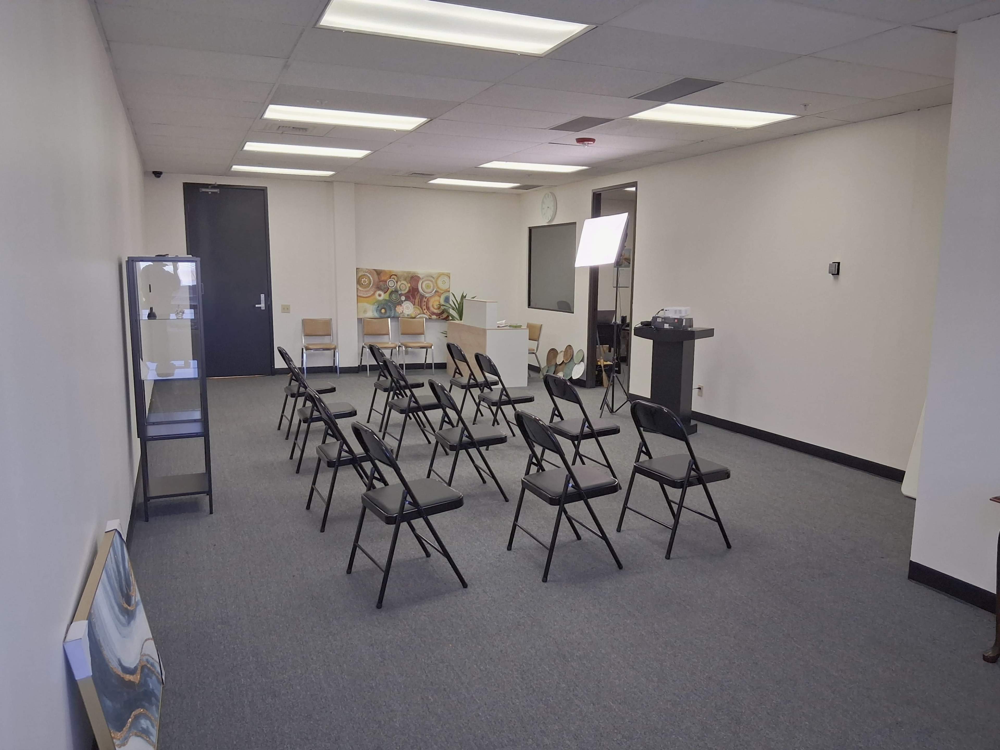 The image shows a spacious room with several black folding chairs arranged in rows, a TV screen on the wall, and a light setup.