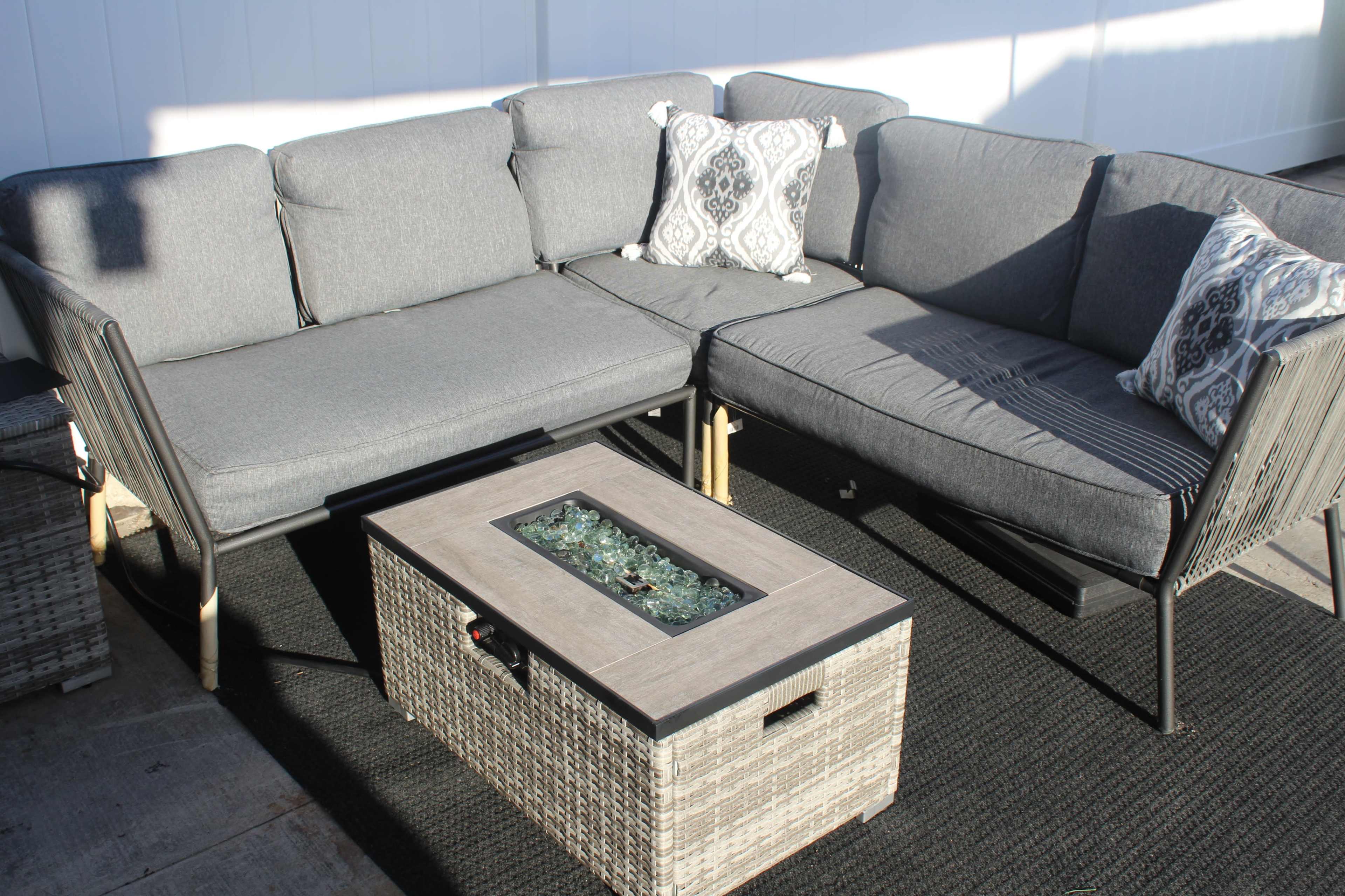 A grey sectional outdoor sofa with a decorative pillow and a rectangular coffee table featuring a glass top surrounded by pebbles.