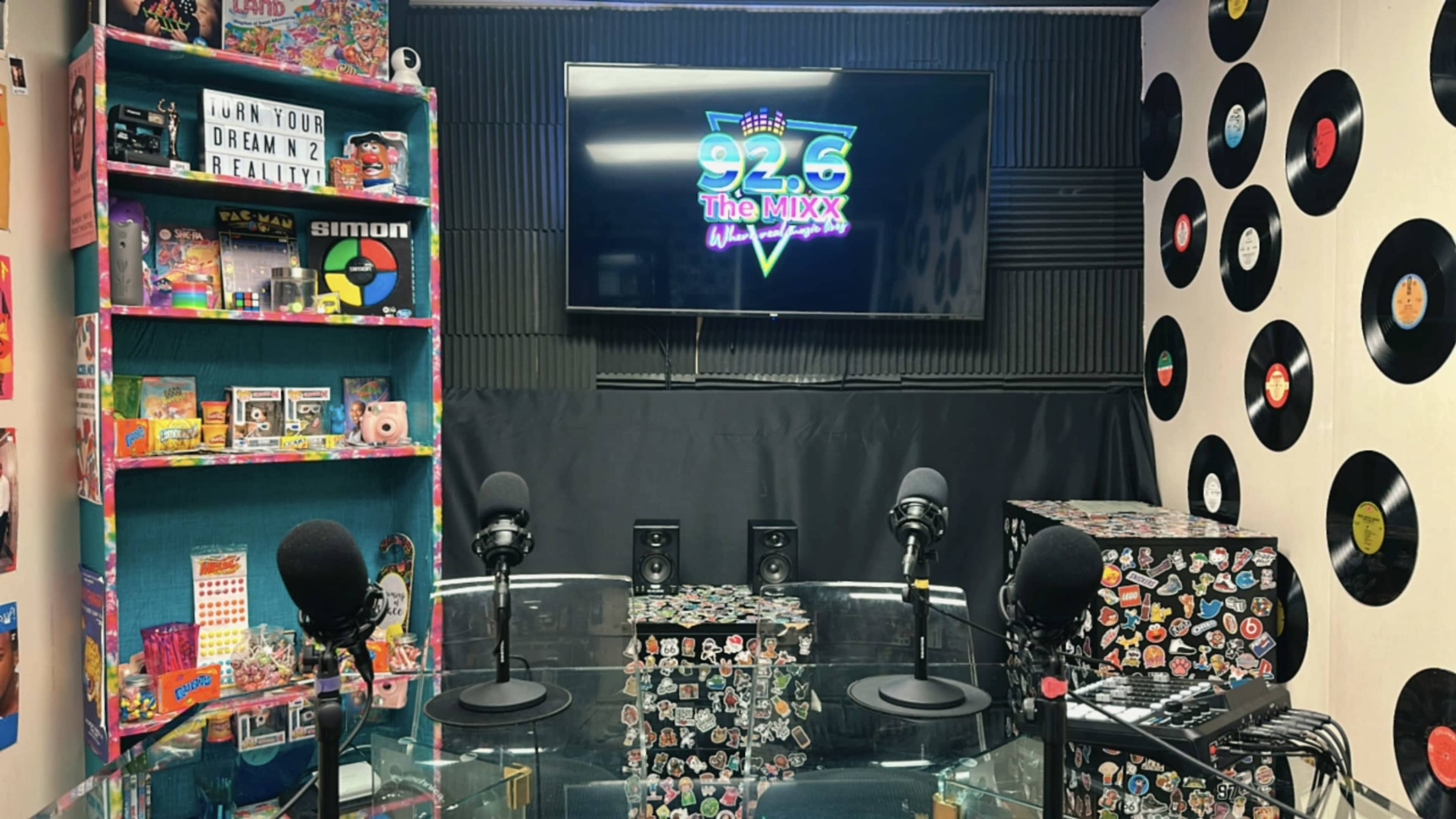 The image shows a colorful radio studio setup with microphones, a large screen displaying the logo "92.6 The MIXX," and shelves filled with various toys and memorabilia.