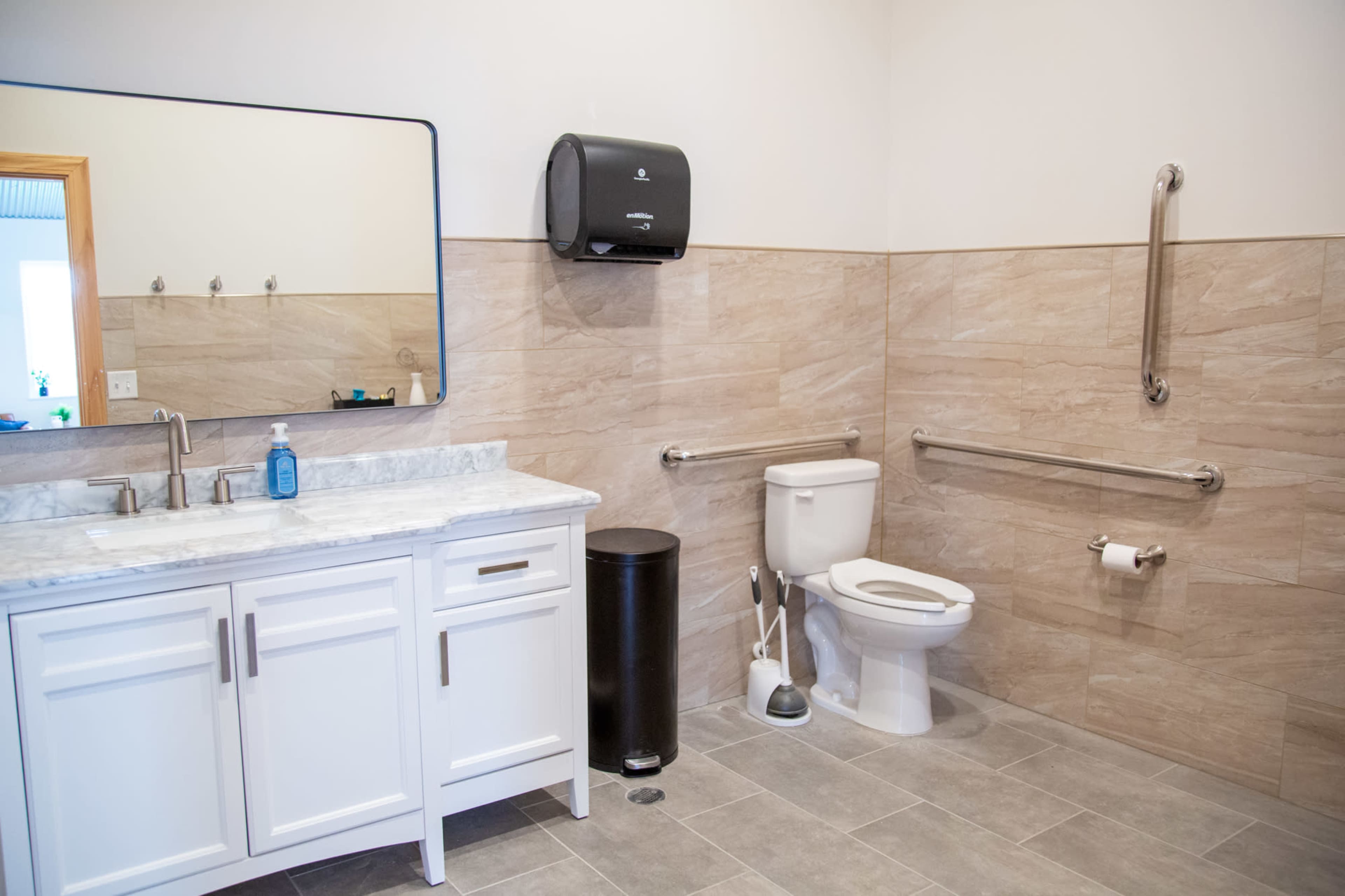 A clean bathroom with a white vanity, a mirror, a toilet with grab bars, a black trash can, and a soap dispenser on the counter.