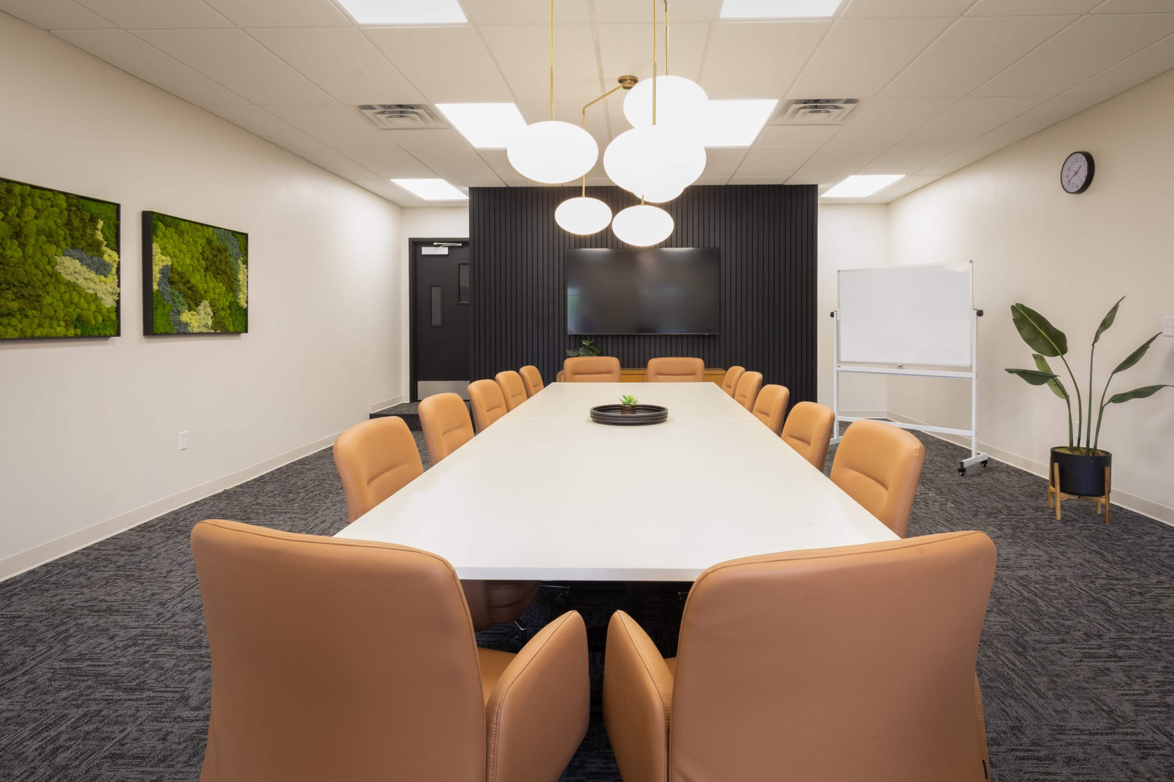 A modern conference room features a long white table surrounded by brown leather chairs, with a wall-mounted screen and decorative plants.