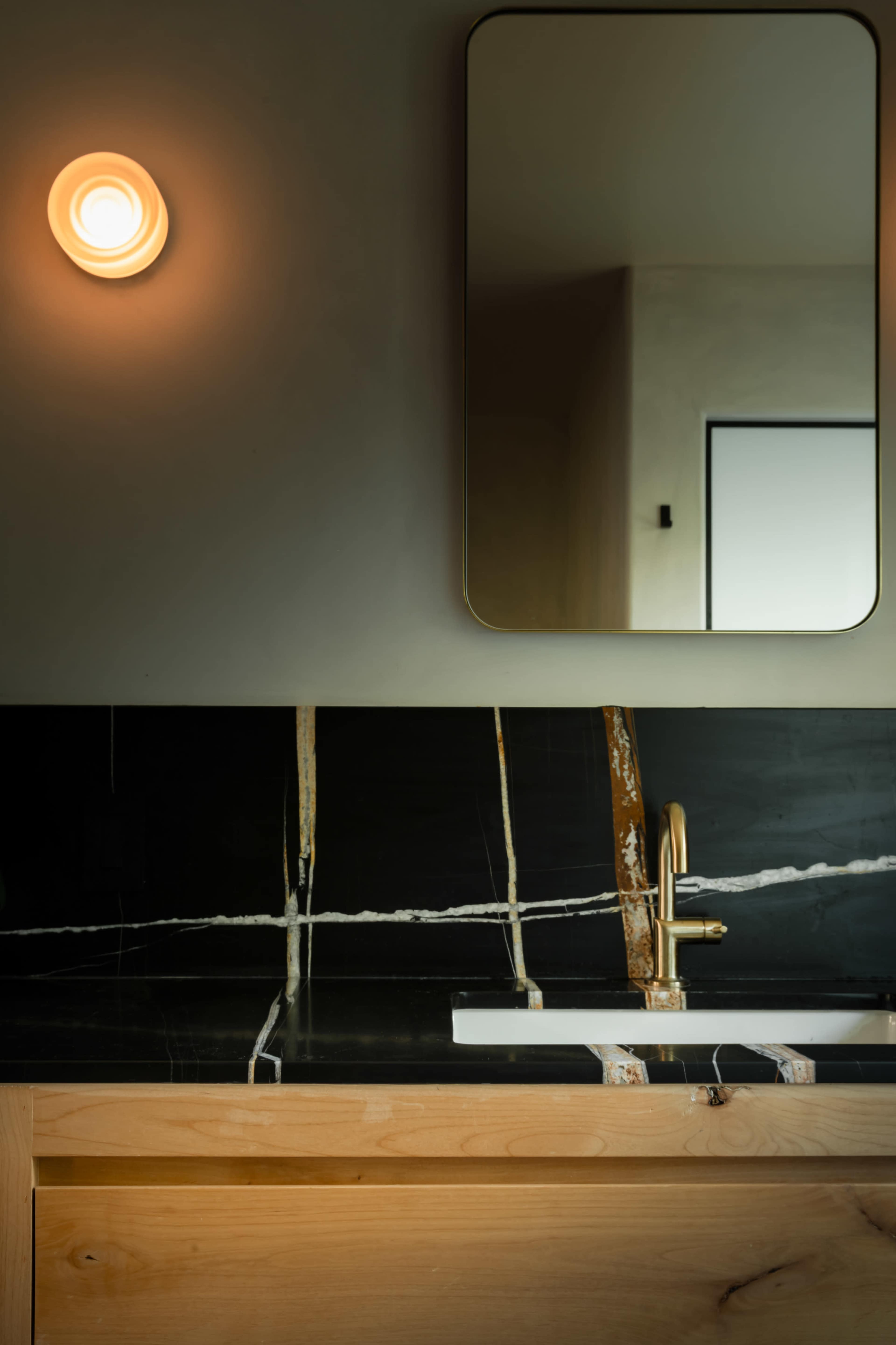 A modern bathroom features a minimalist wooden vanity with a striking black countertop and a round wall sconce illuminating a rectangular mirror.