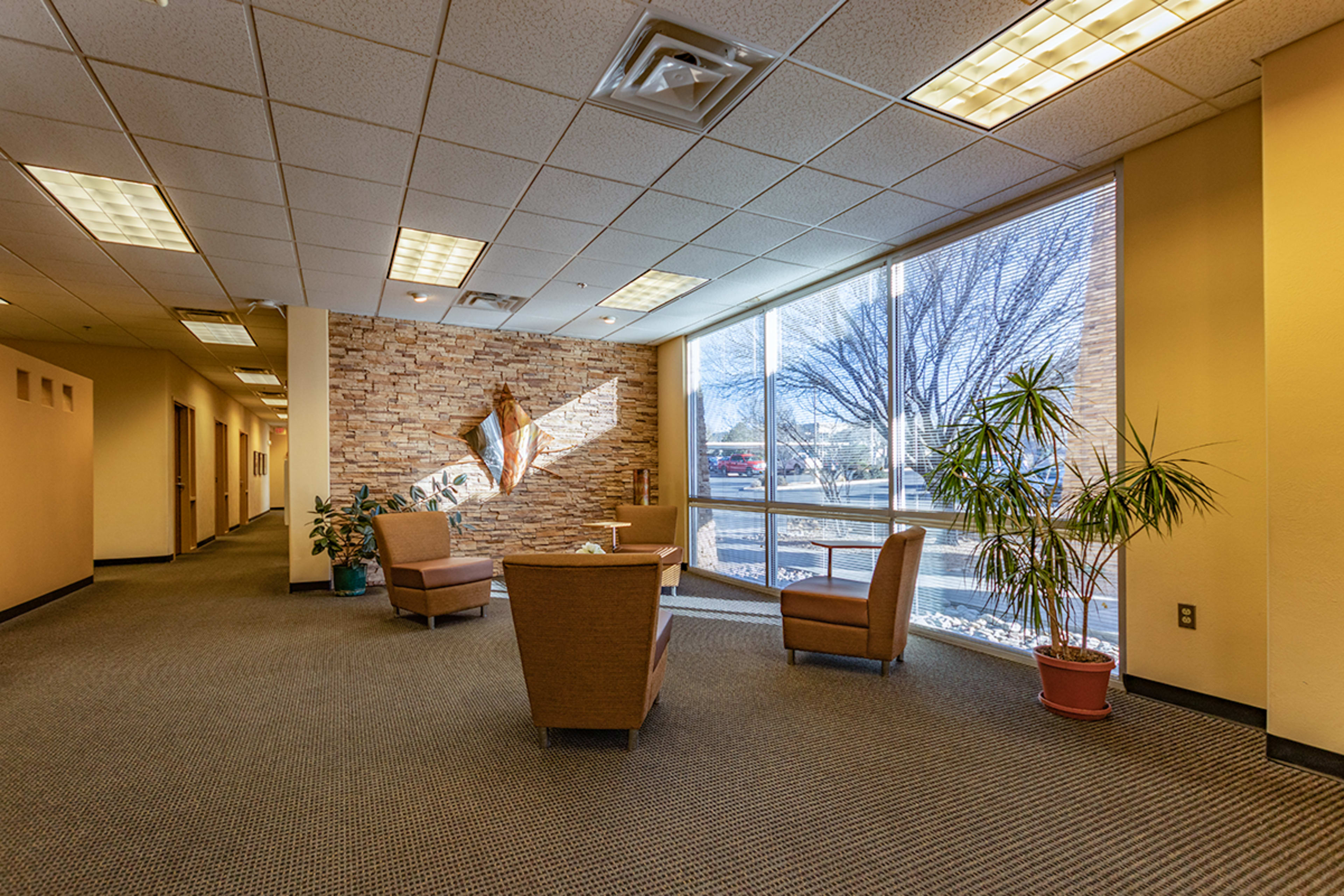 A seating area with two chairs and a potted plant is located beside large windows in a hallway featuring stone walls.