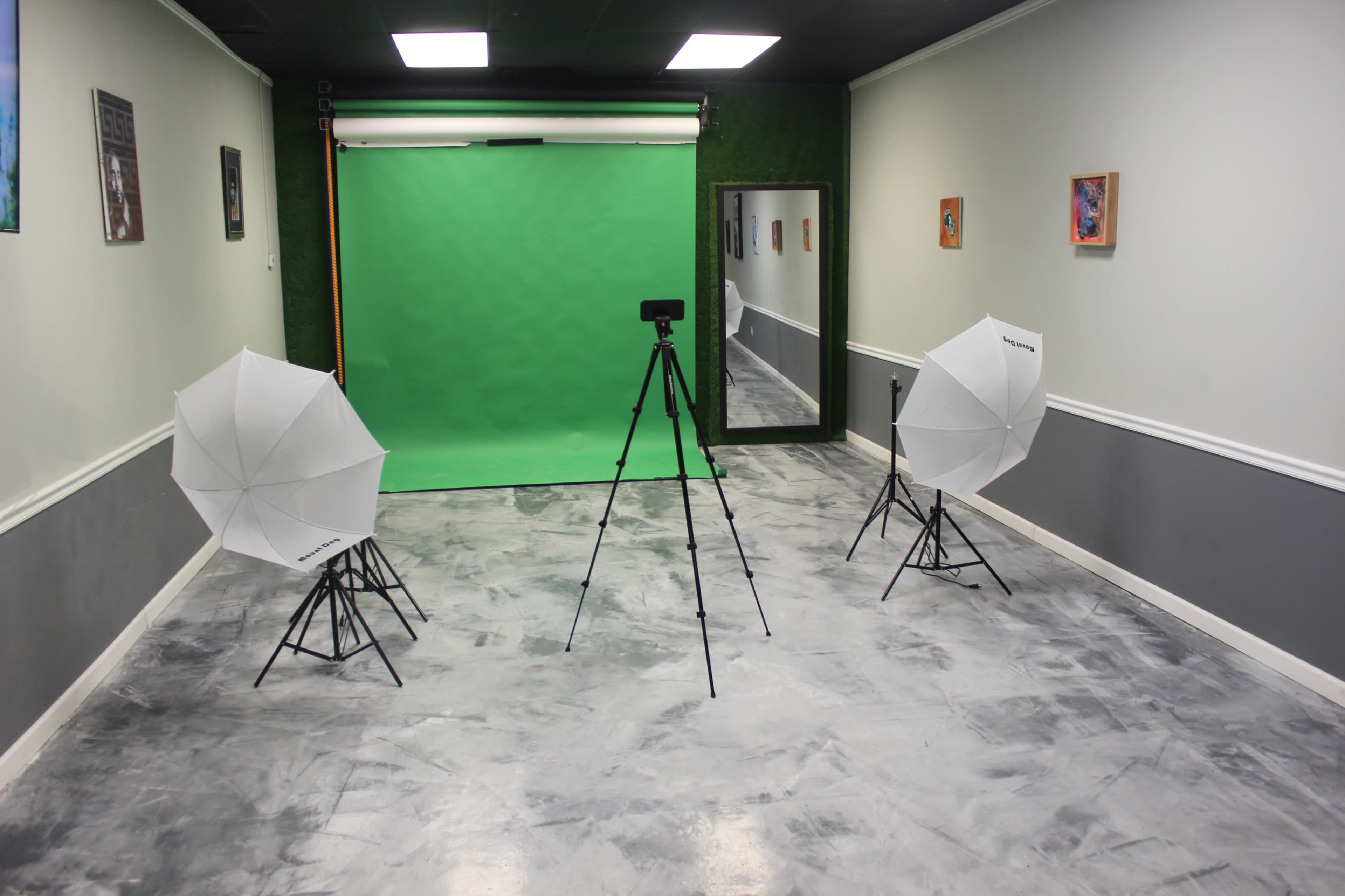 The image shows a photography studio setup with a green backdrop, two softbox lights on tripods, and a camera on a tripod in a sparsely decorated room.