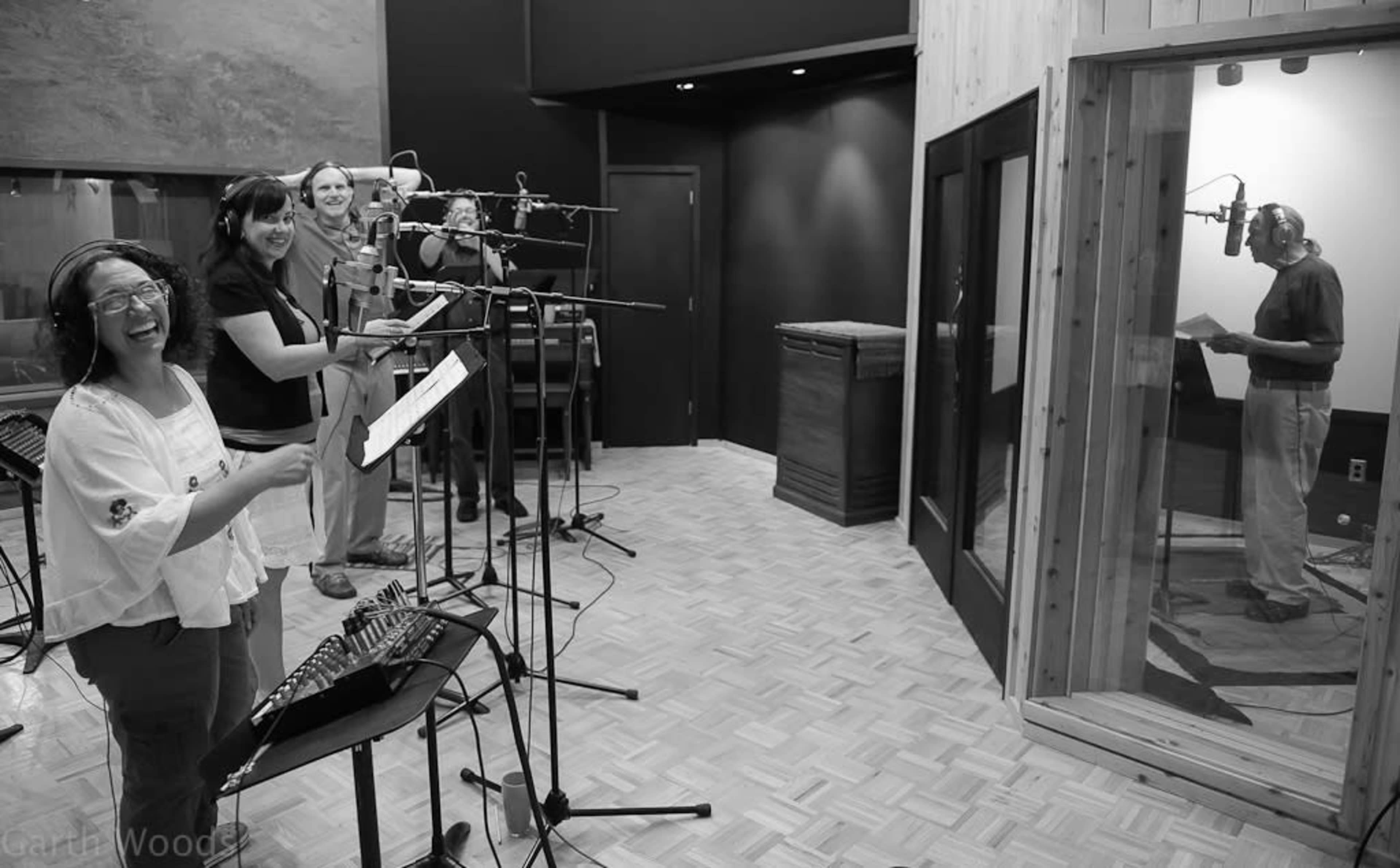 Musicians perform vocals in a recording studio while one singer records in a separate booth.