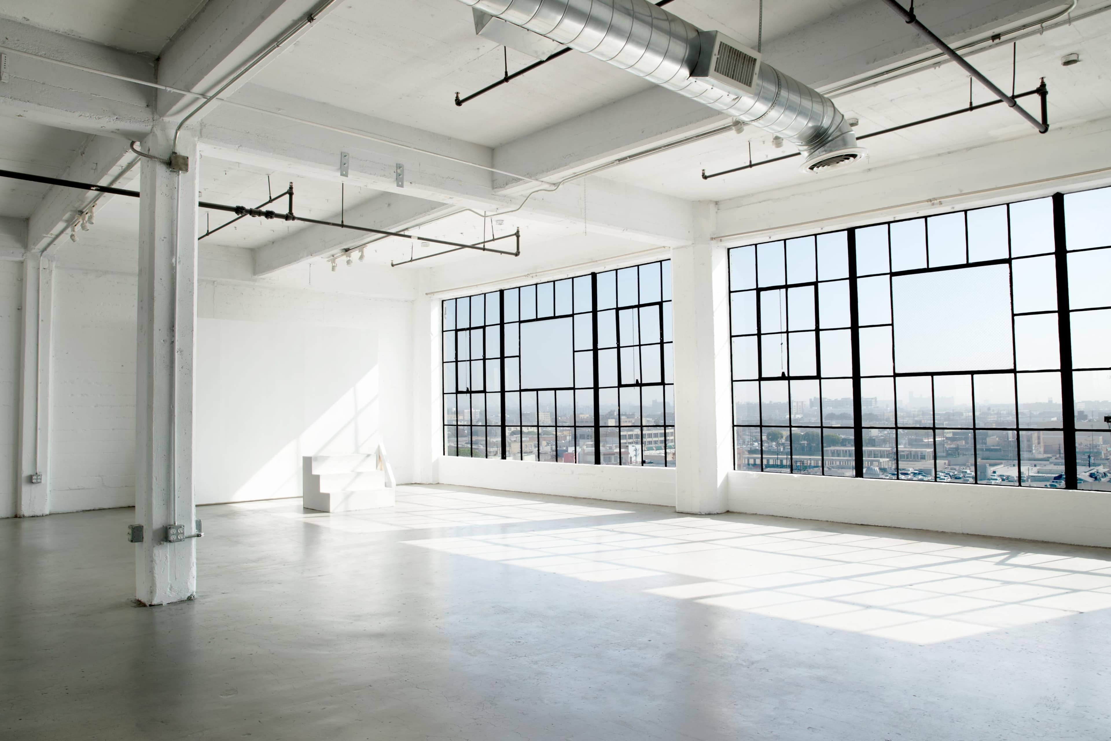 The image shows a spacious, empty room with large windows allowing natural light to fill the space.