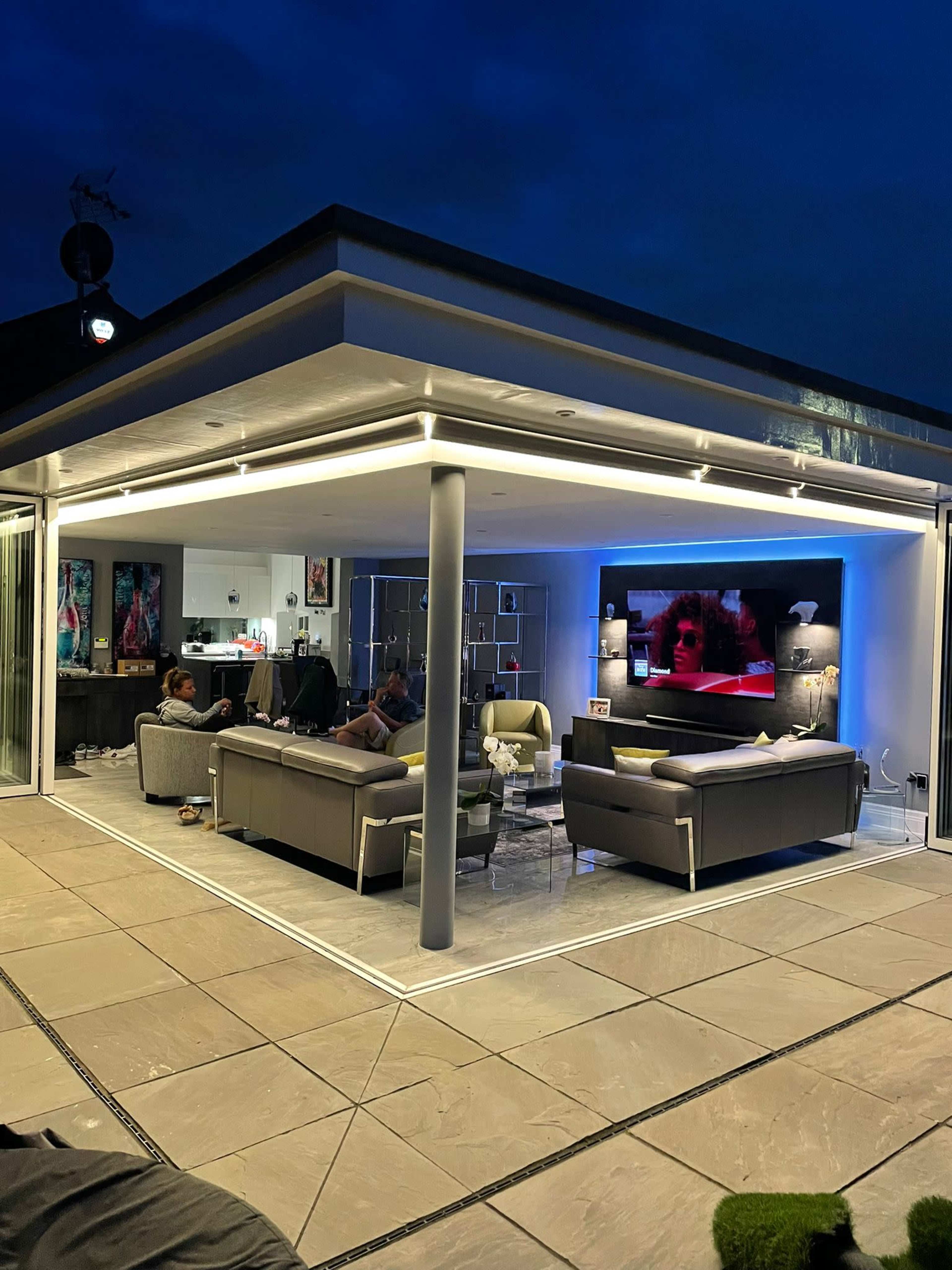 A modern, open-plan living area at night with a large television displaying a scene on one wall and comfortable seating arranged around it.
