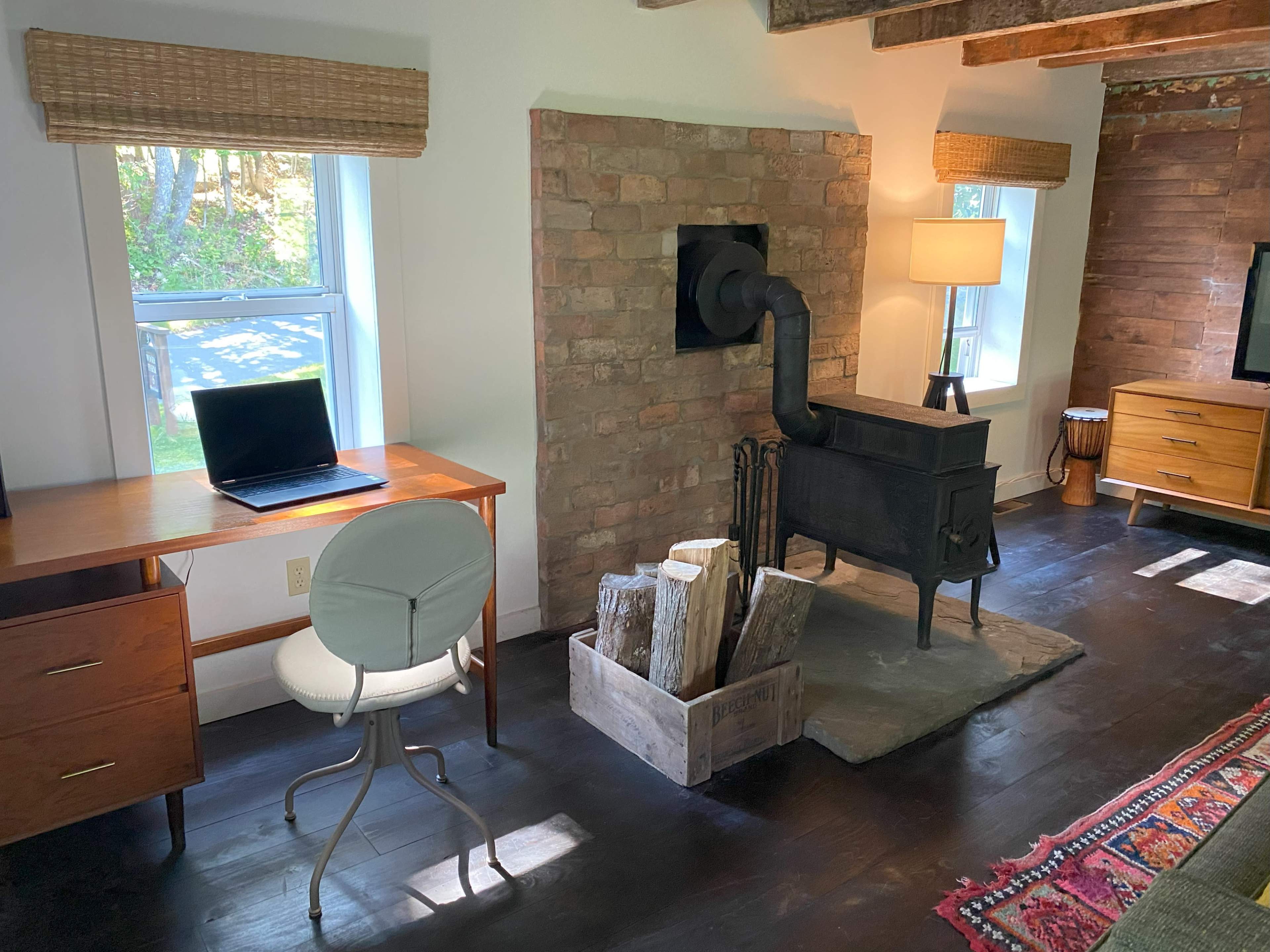 The image shows a cozy workspace featuring a wooden desk with a laptop next to a brick wall that has a black stove, accompanied by a lamp and a crate of logs.