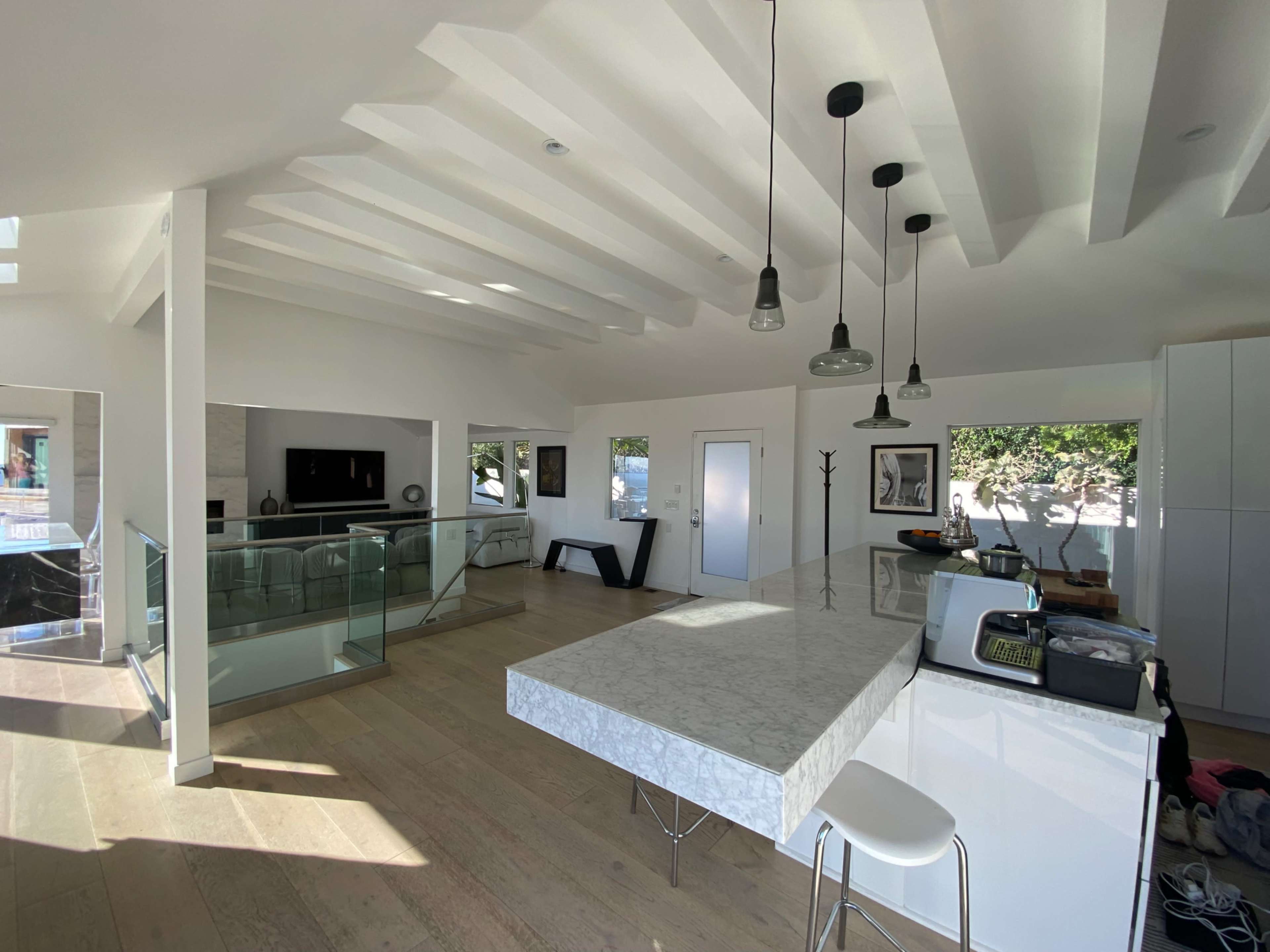 The image shows a modern kitchen and living area with a marble countertop, pendant lights, and large glass windows allowing natural light to enter.