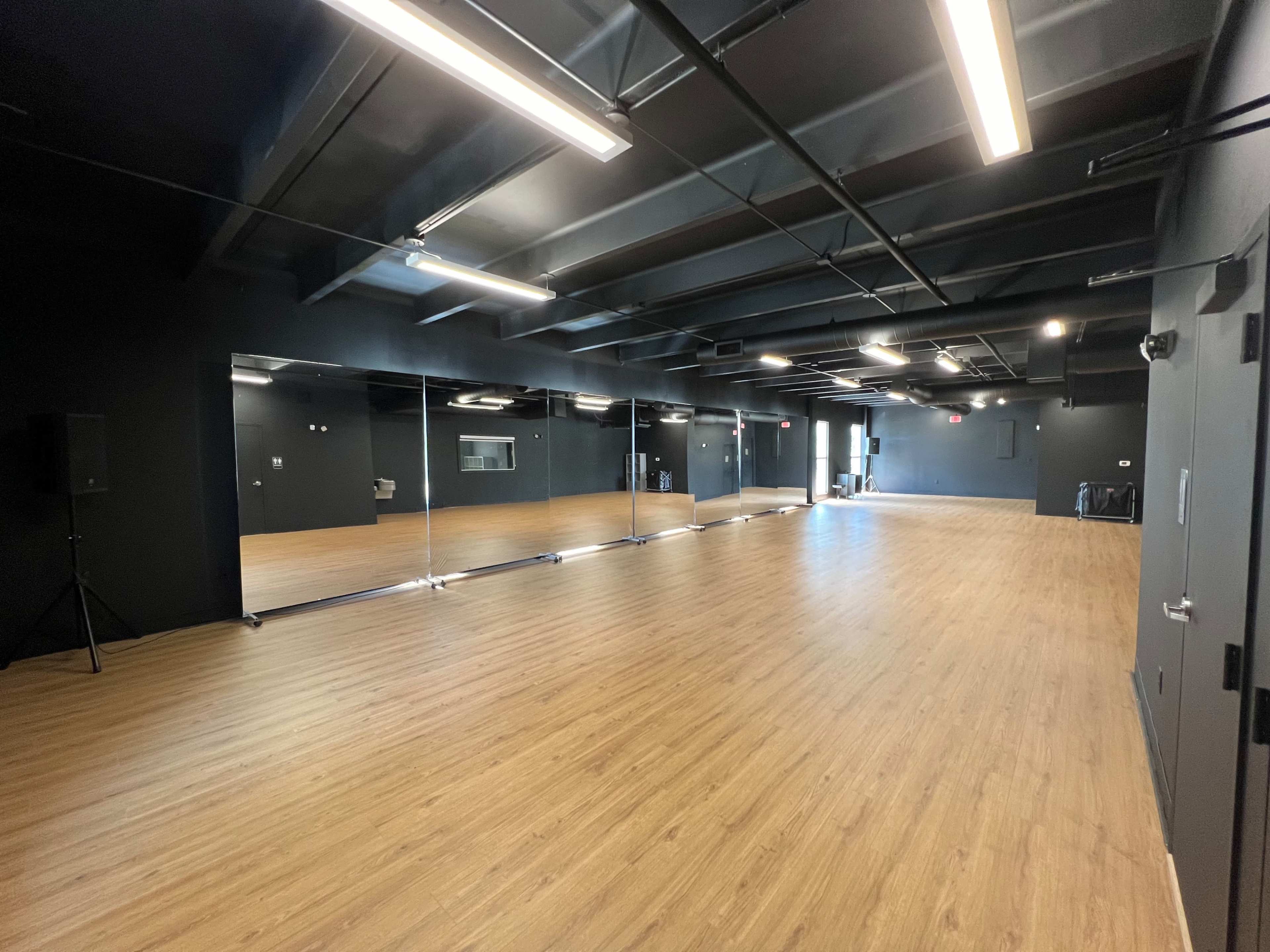 A spacious dance studio with wooden floors and a large mirror wall, illuminated by overhead lighting.