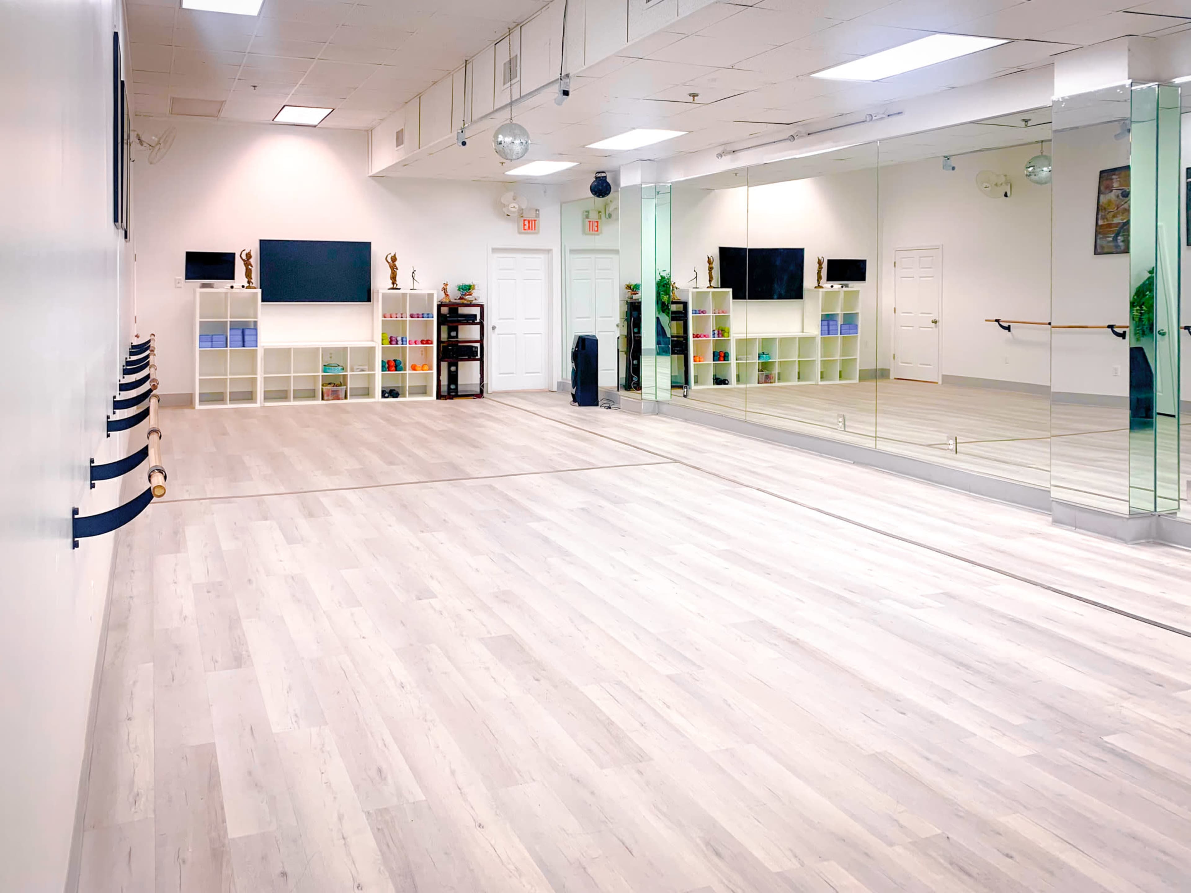 The image shows a spacious, well-lit studio with a wooden floor, mirrors on one wall, and shelves displaying various items.