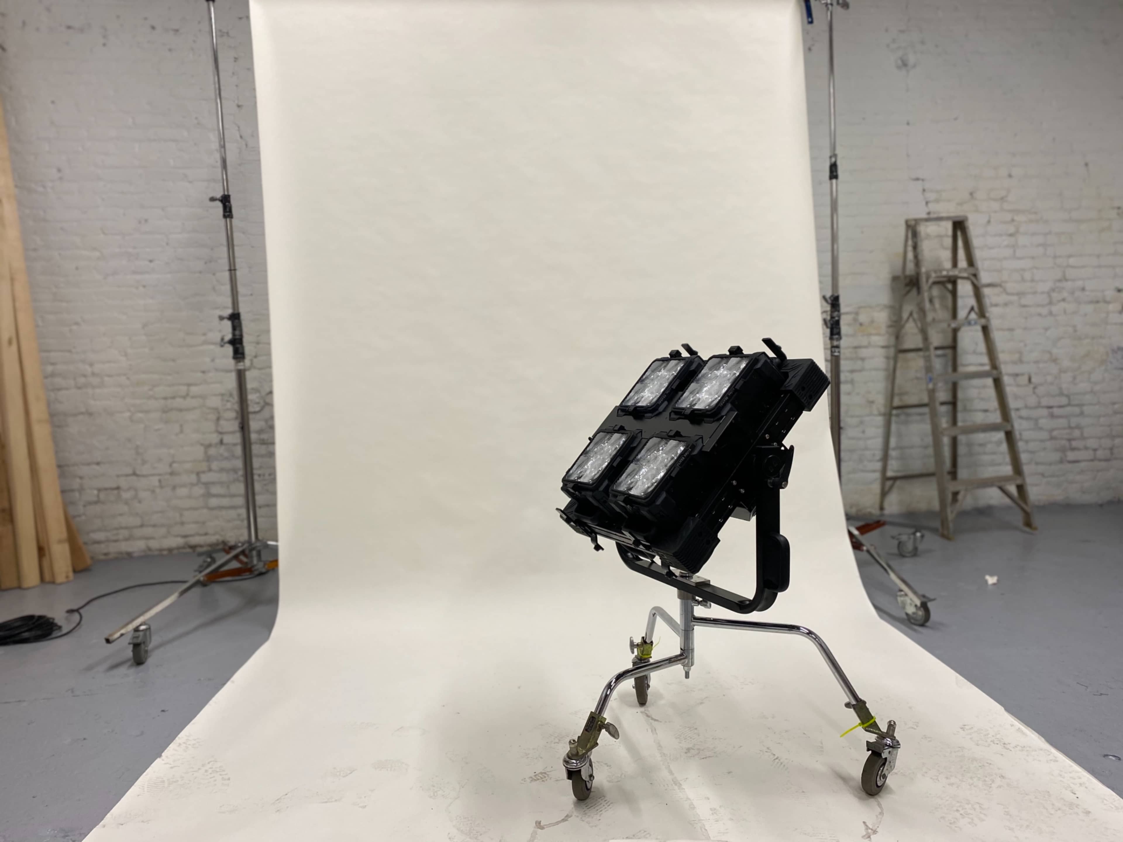 A lighting setup on a stand is positioned in front of a plain backdrop in a studio space.