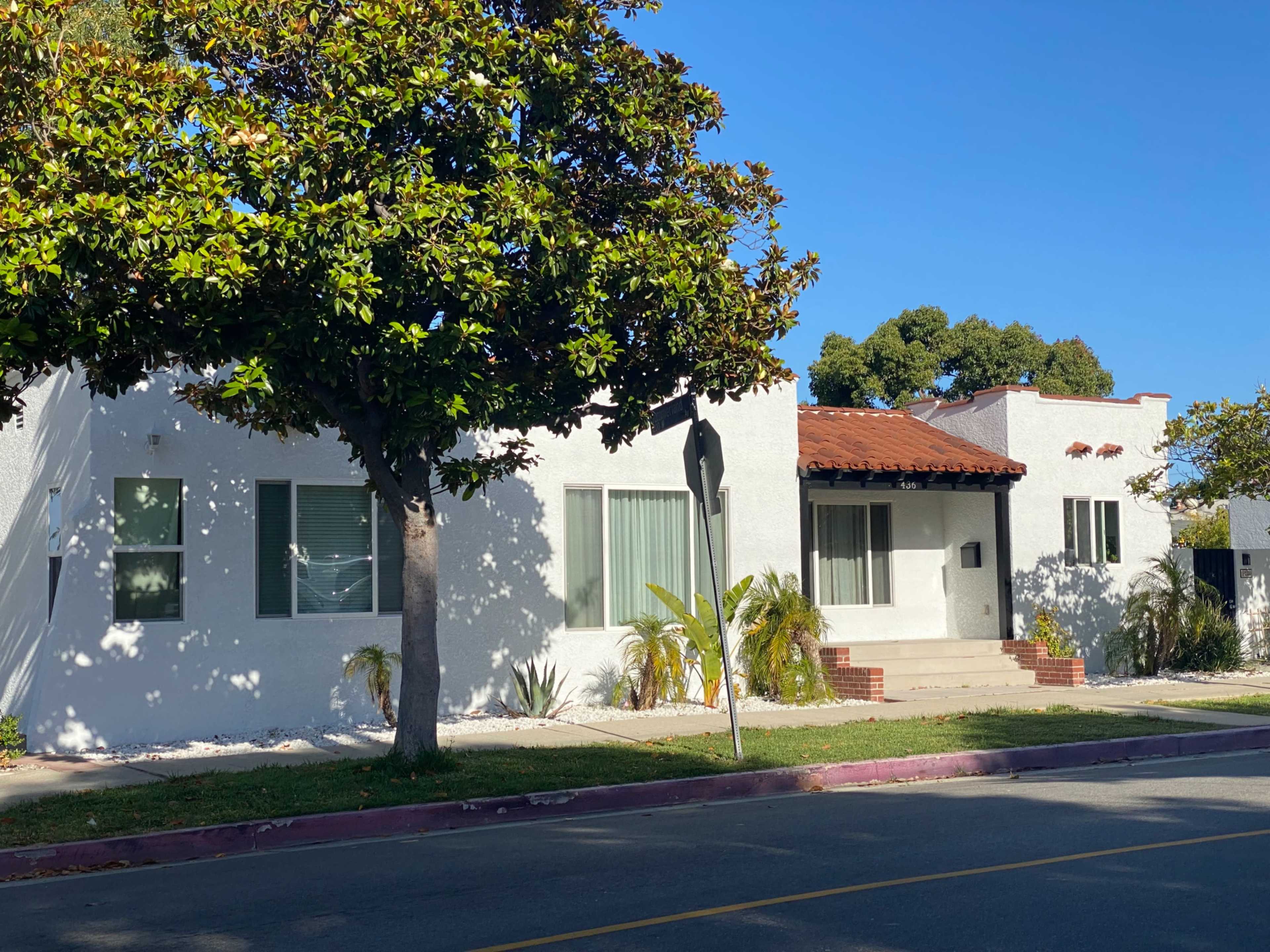 A white residential building with a red-tiled roof, situated on a sunny street lined with trees.