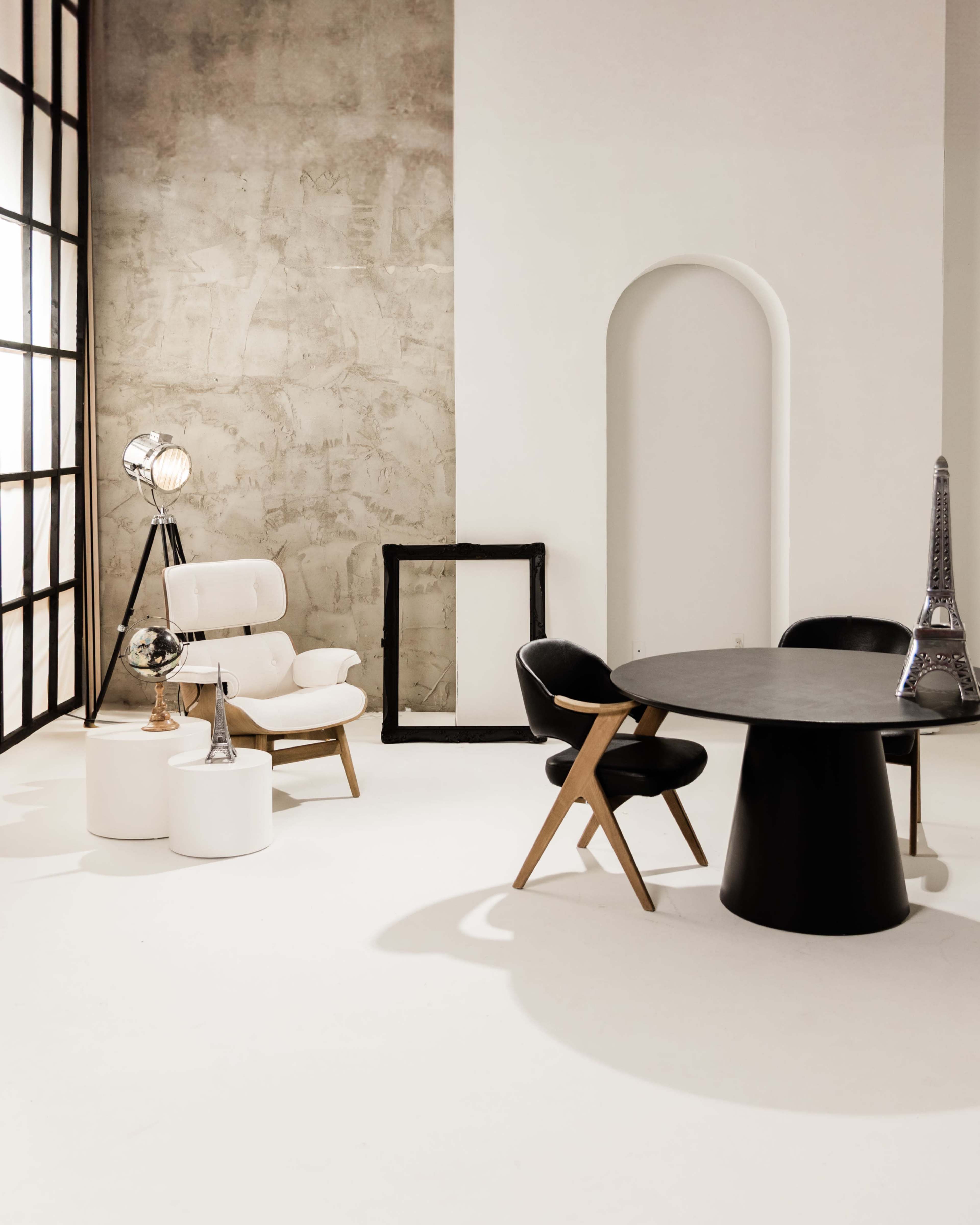 The image features a minimalist interior with a round black dining table, two contrasting chairs, a white armchair, and a decorative Eiffel Tower model against a textured wall.