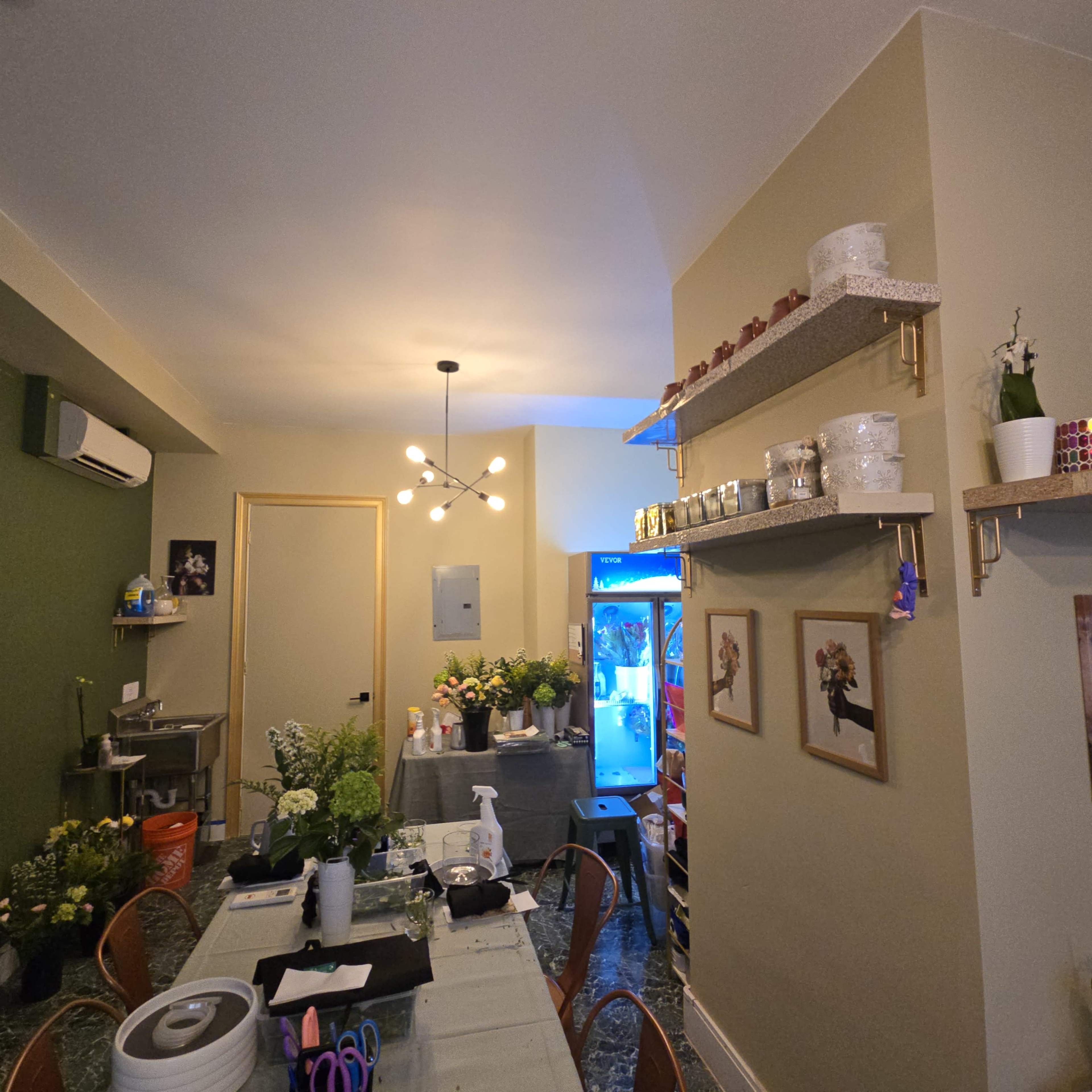 The image shows a well-decorated room featuring a table with various floral arrangement materials, a refrigerator in the corner, and decorative shelves displaying plants and supplies on the walls.