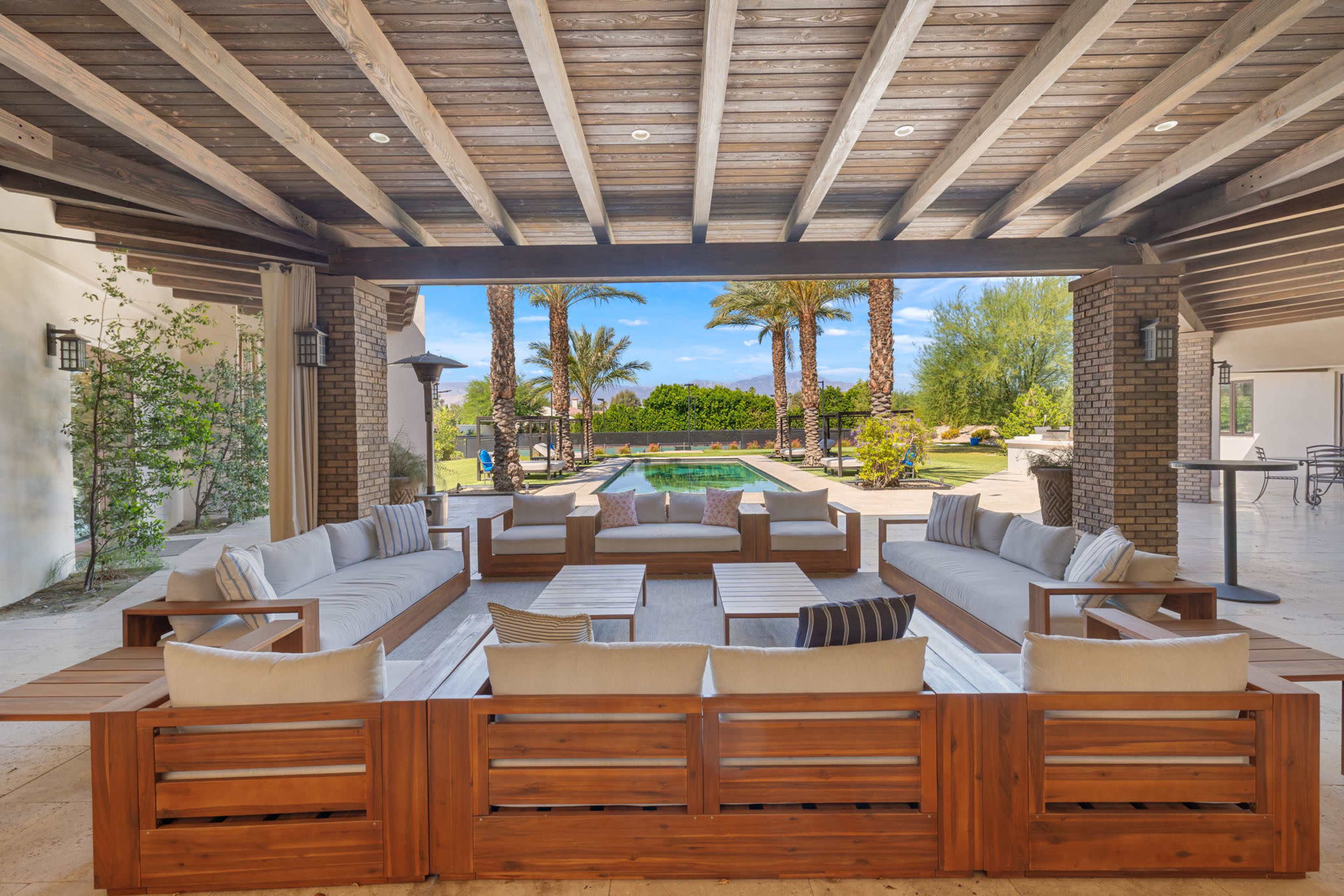 The image shows a covered outdoor seating area with large sofas and coffee tables, overlooking a pool surrounded by palm trees and landscaped greenery.