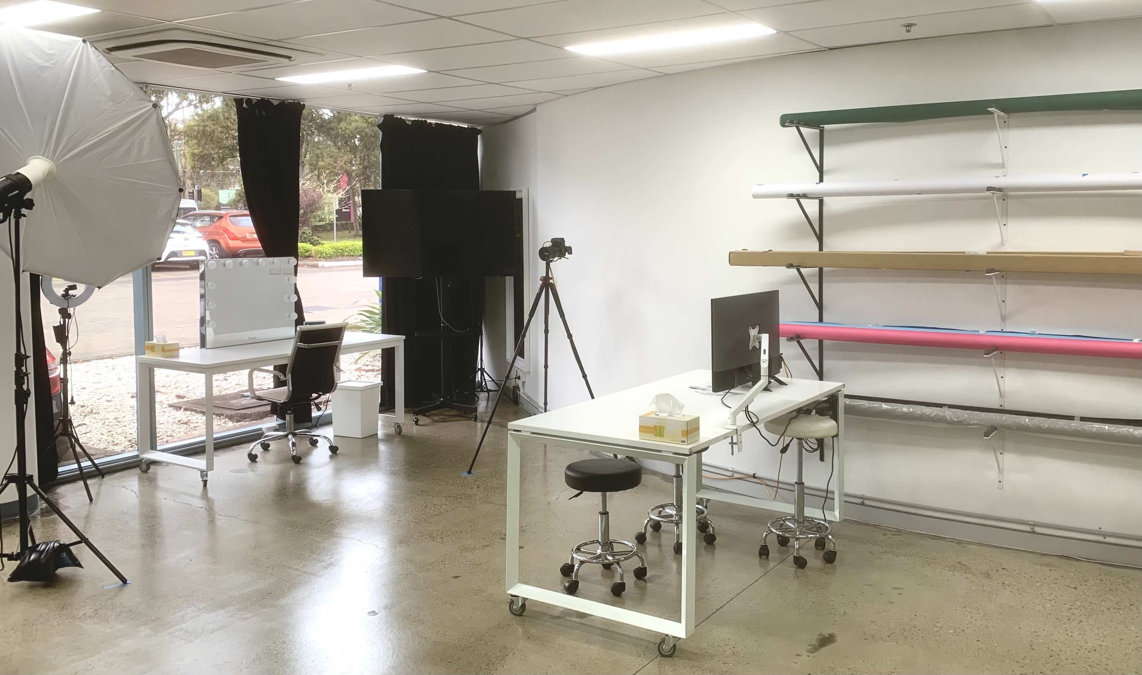 A bright photography studio with two white desks, a backdrop rack with colorful paper, and equipment including a camera and lighting setup.