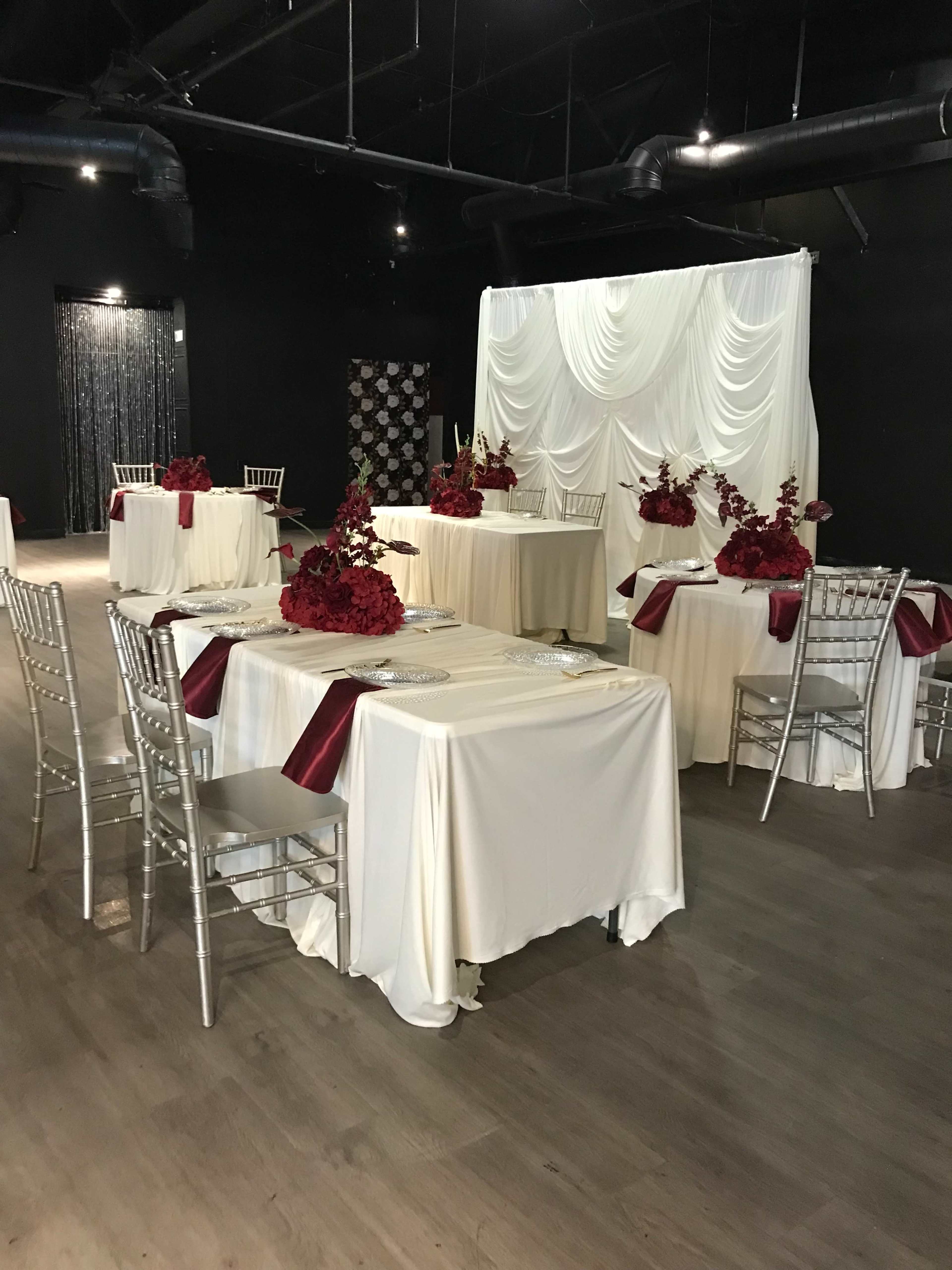 The image shows a banquet hall set with several covered tables adorned with white tablecloths and red floral centerpieces.