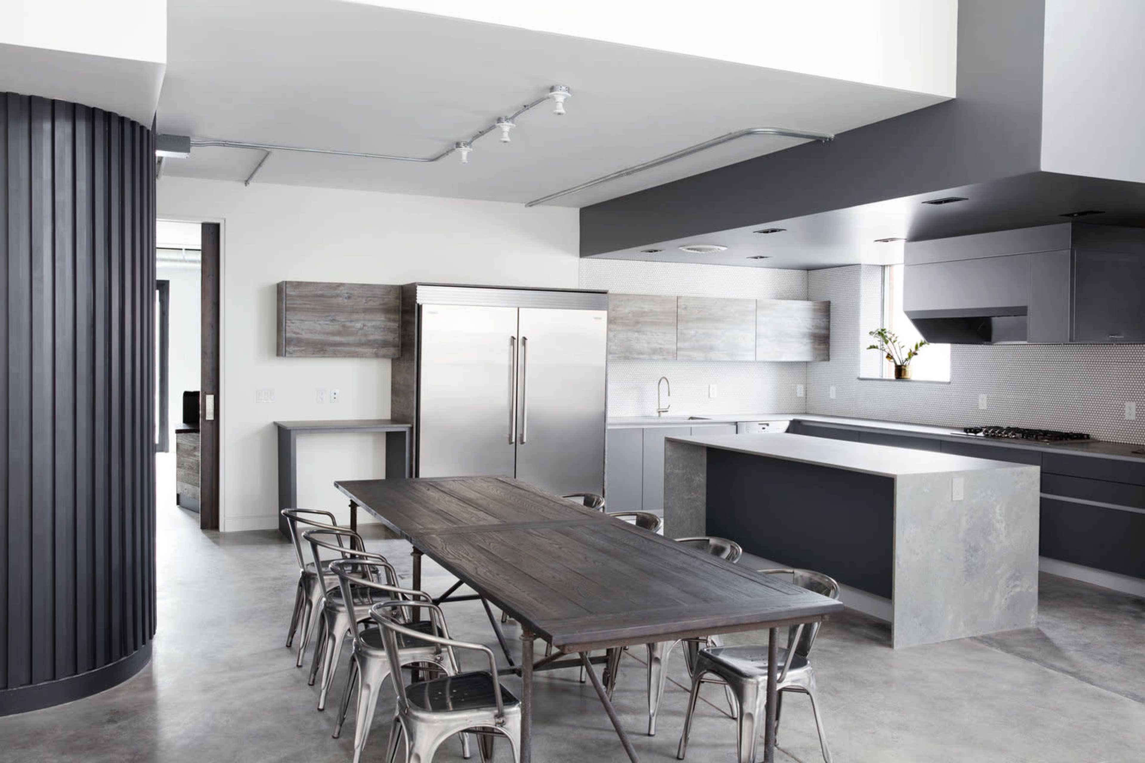 A modern kitchen features stainless steel appliances, gray cabinetry, and a long wooden dining table surrounded by metal chairs.
