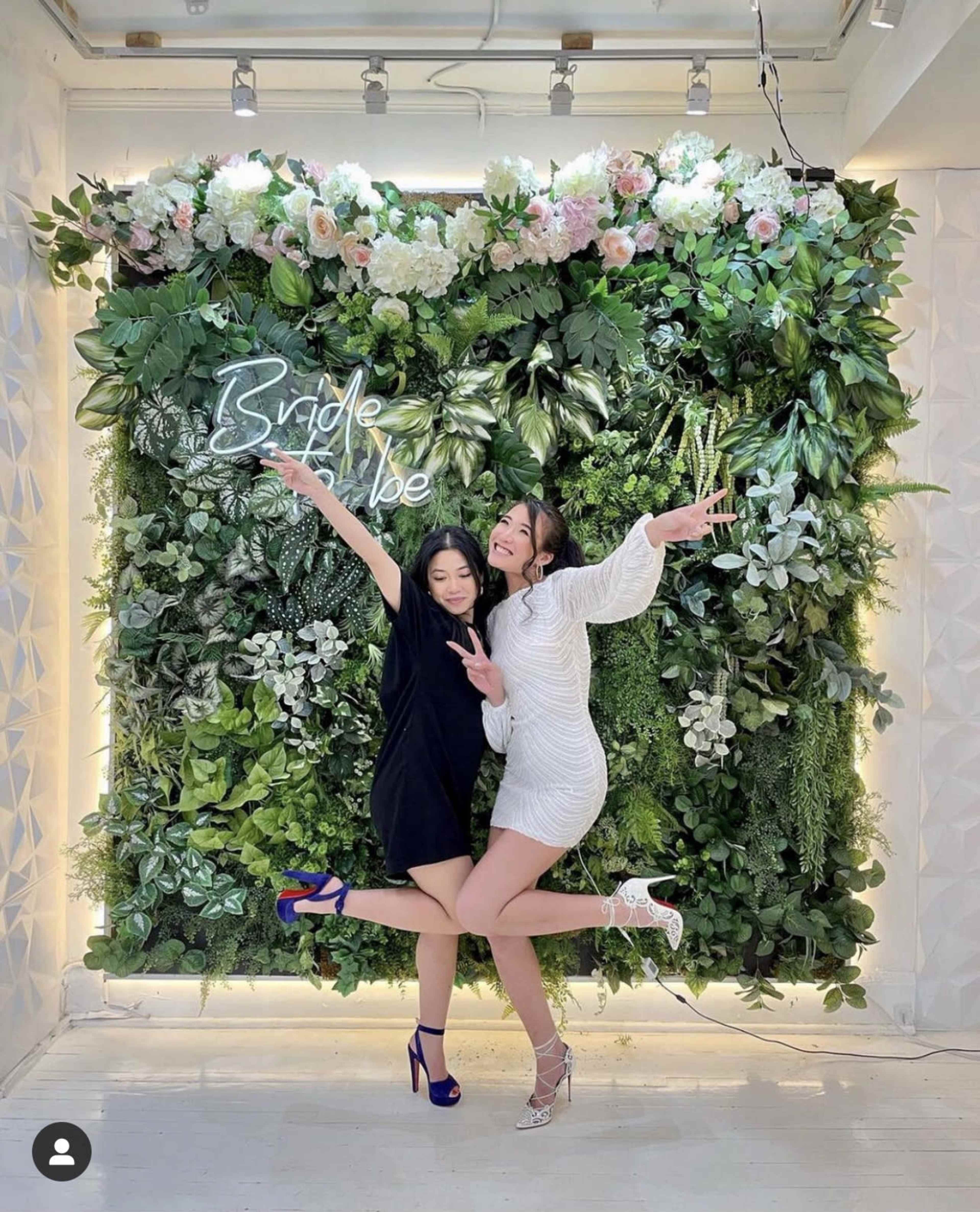 Two women pose playfully in front of a green floral backdrop that features the words "Bride to Be."