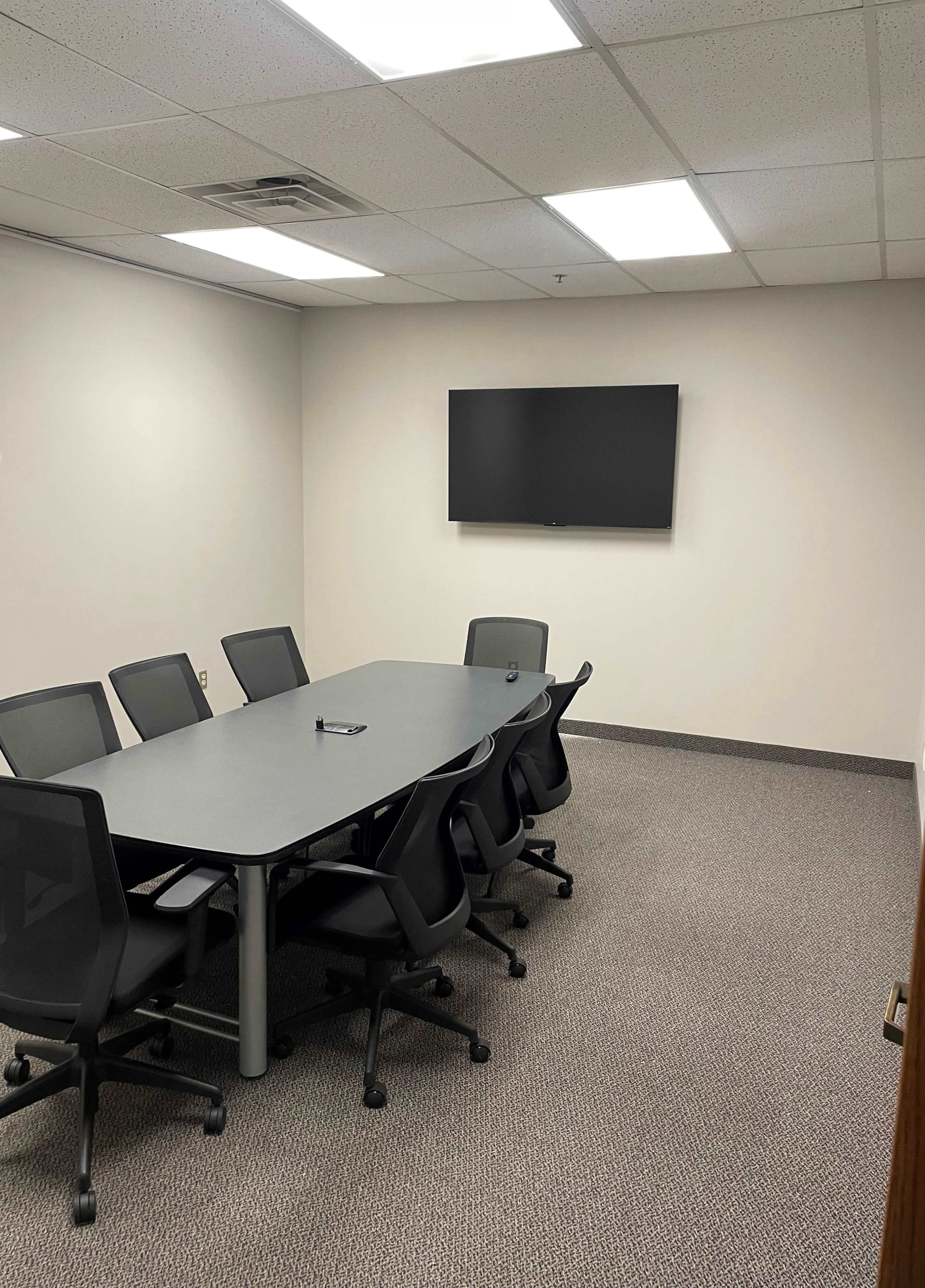 A conference room features a rectangular table surrounded by six swivel chairs and a wall-mounted flat-screen TV.