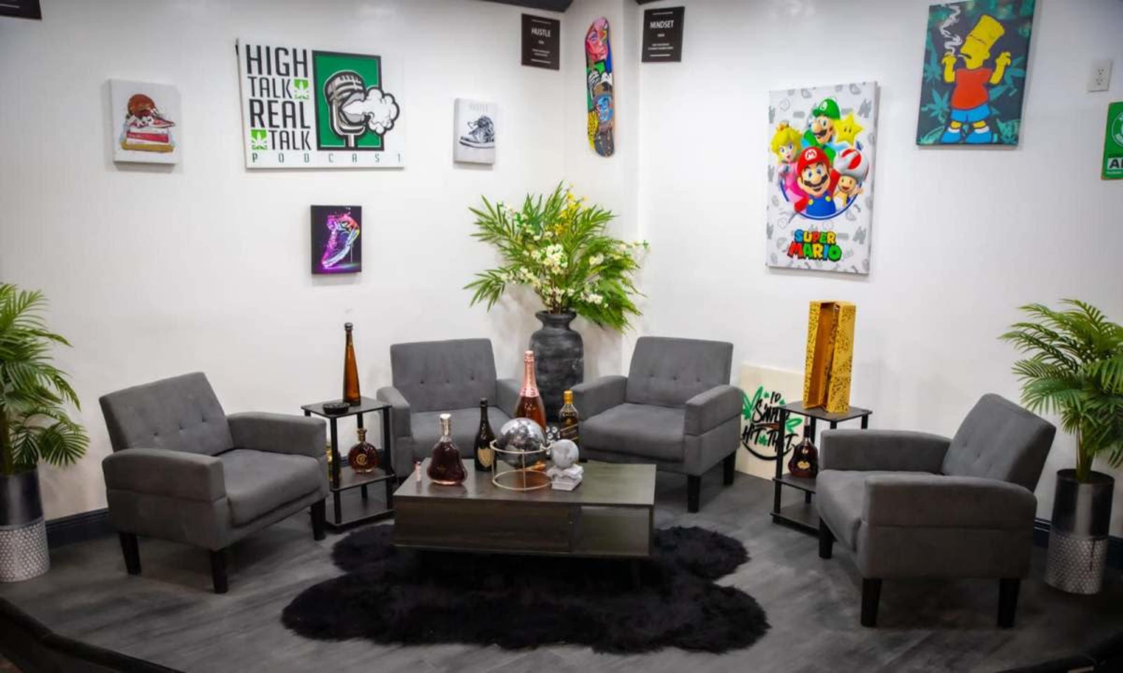 The image shows a cozy podcast recording studio featuring gray chairs arranged around a coffee table, with decorative plants and colorful artwork on the walls.