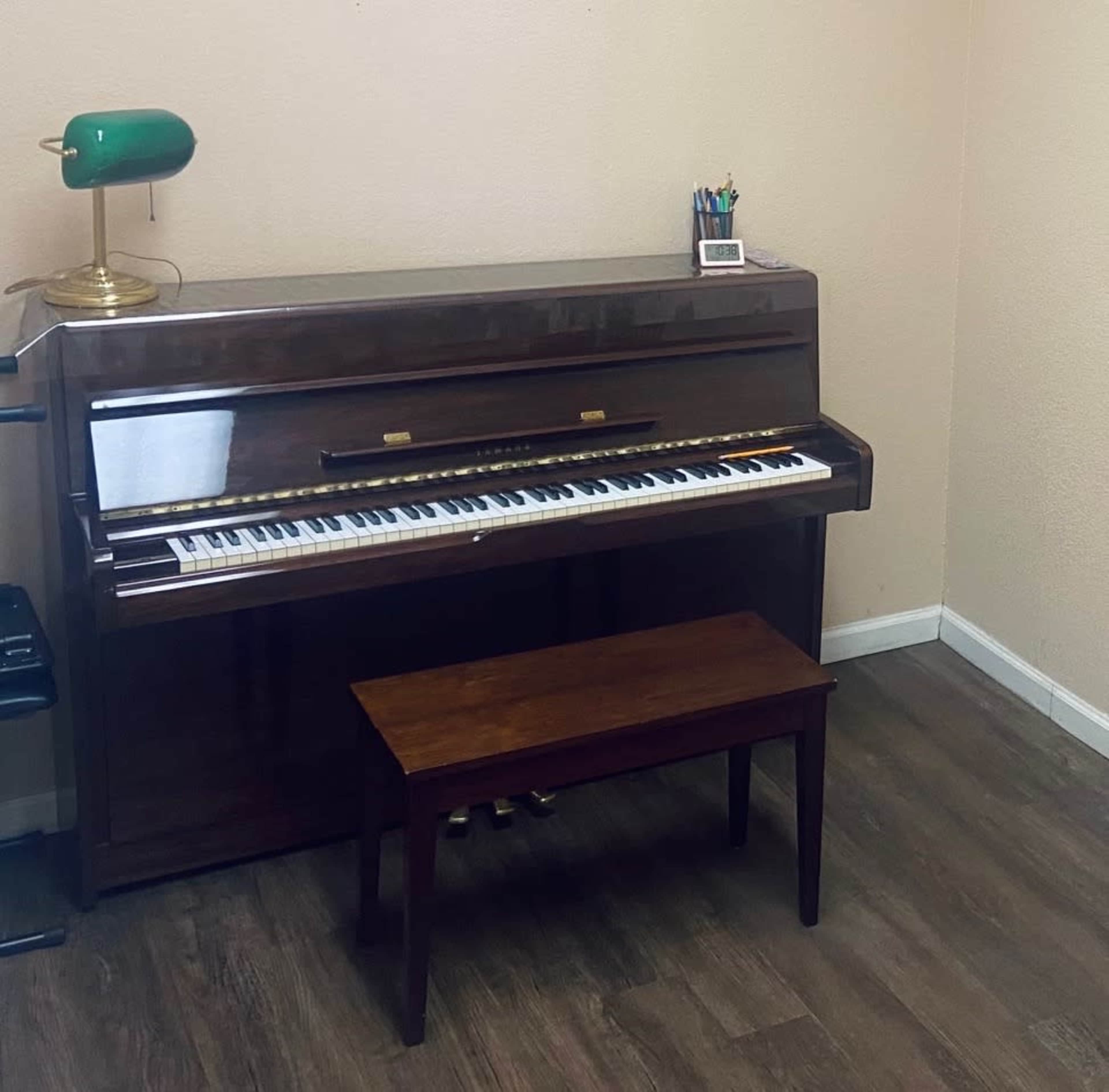 A wooden piano with a matching bench sits against a wall in a room, accompanied by a green desk lamp and a collection of pens on the side.