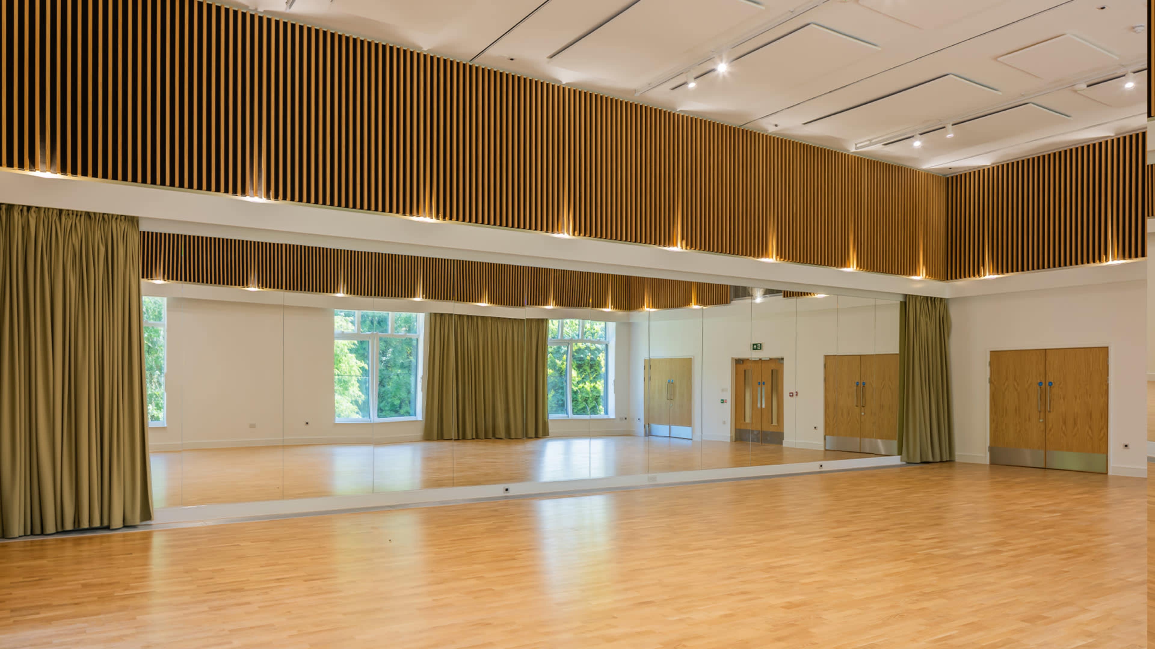 A spacious room with wooden paneling, large mirrors, and natural light coming through windows, featuring a wooden floor and green curtains.