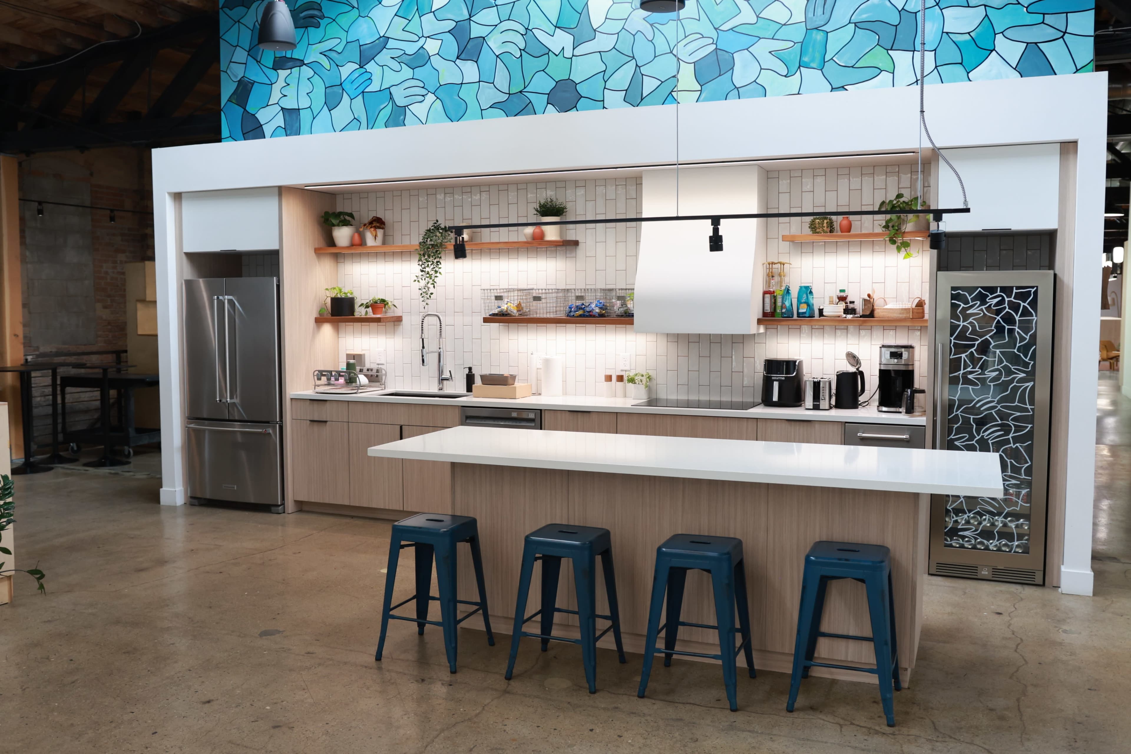 The image shows a modern kitchen with a blue tile backsplash, wooden cabinetry, and several bar stools arranged in front of a long countertop.