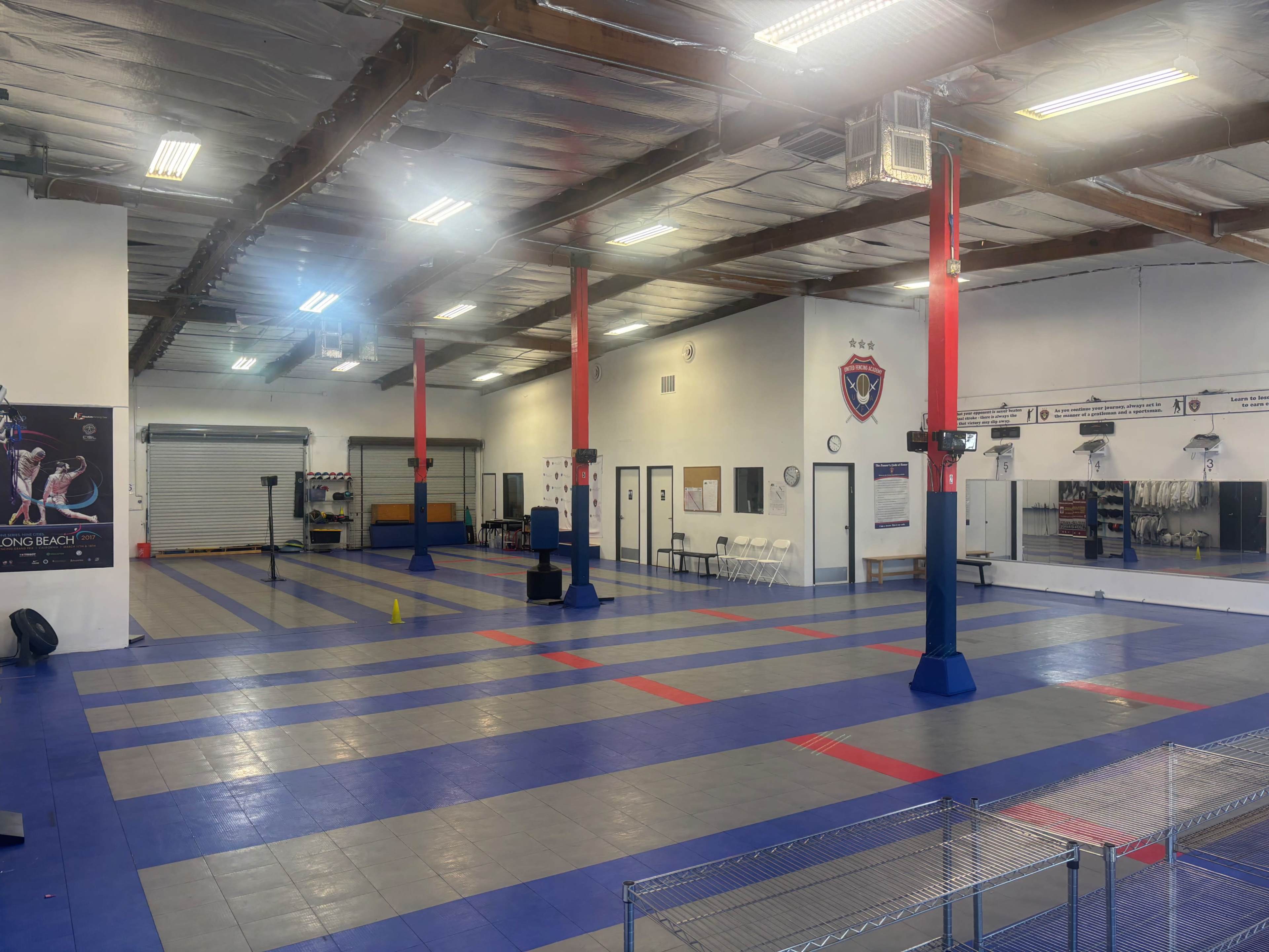 The image shows an indoor martial arts training facility featuring a large open space with blue and gray mats, red pillars, and various training equipment.