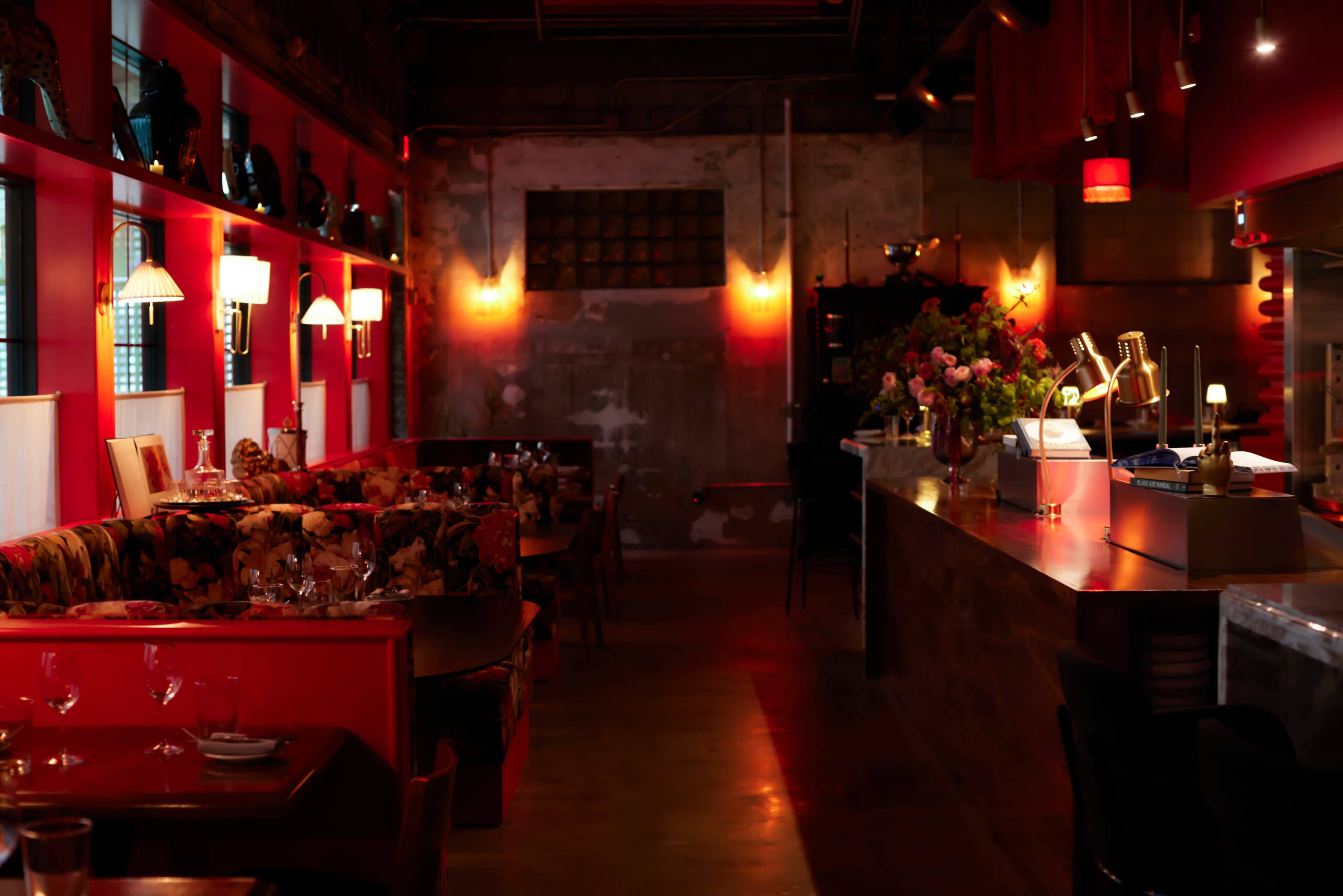 The image shows a dimly lit restaurant interior featuring red walls, floral-patterned seating, and a bar area with decorative lighting.