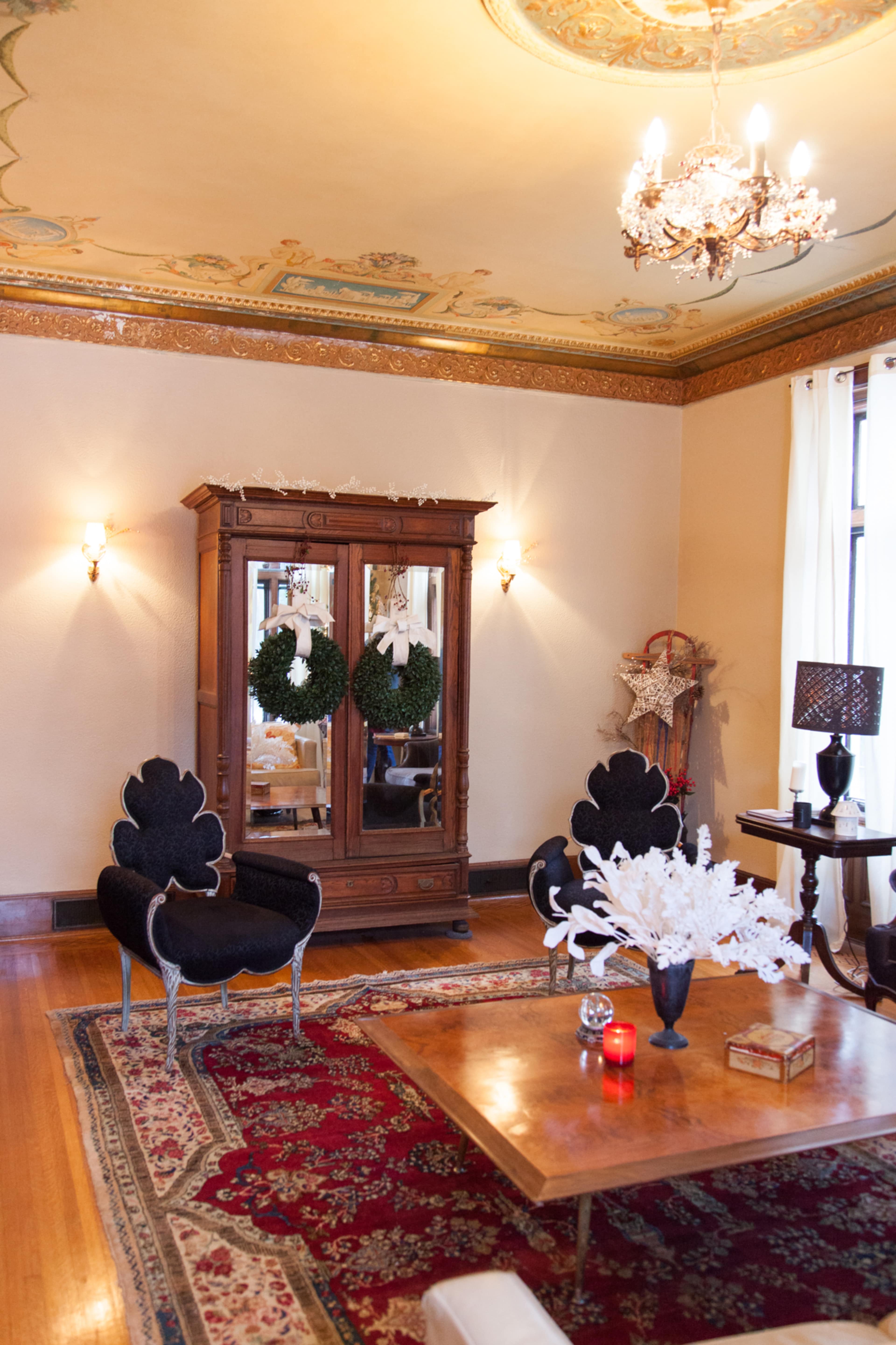 The image shows a cozy living room featuring ornate furnishings, a decorative area rug, and a vintage cabinet adorned with Christmas wreaths.