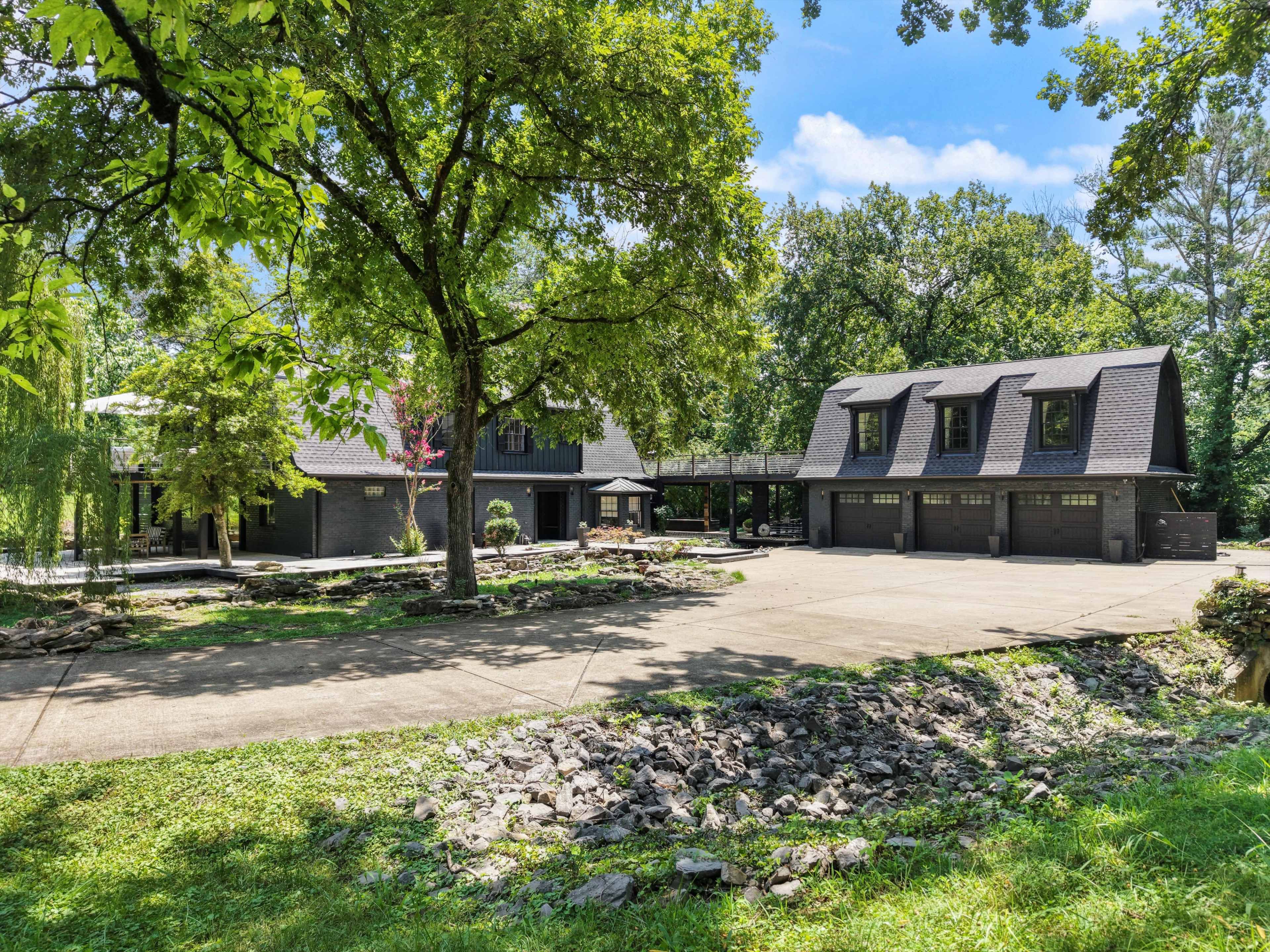 The image shows a large, modern home with multiple garage doors and a wide driveway, surrounded by trees and landscaping.