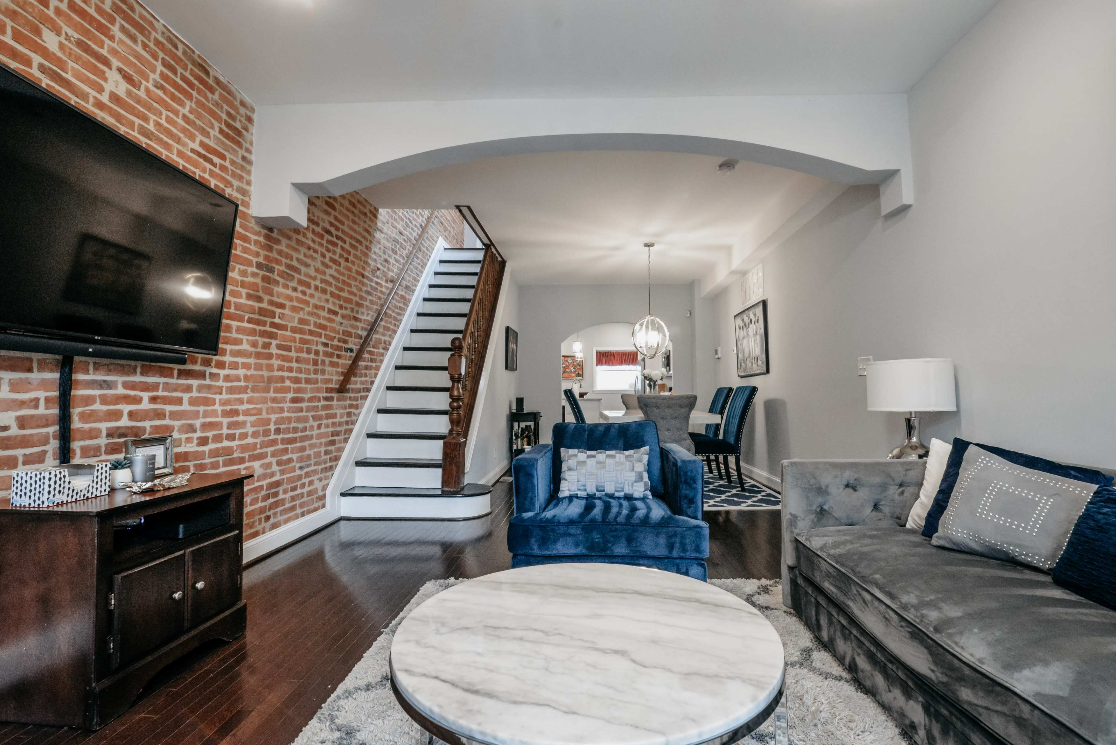 A living room featuring a blue sofa, a marble coffee table, a large television, and an exposed brick wall, with a staircase leading to an upper level in the background.