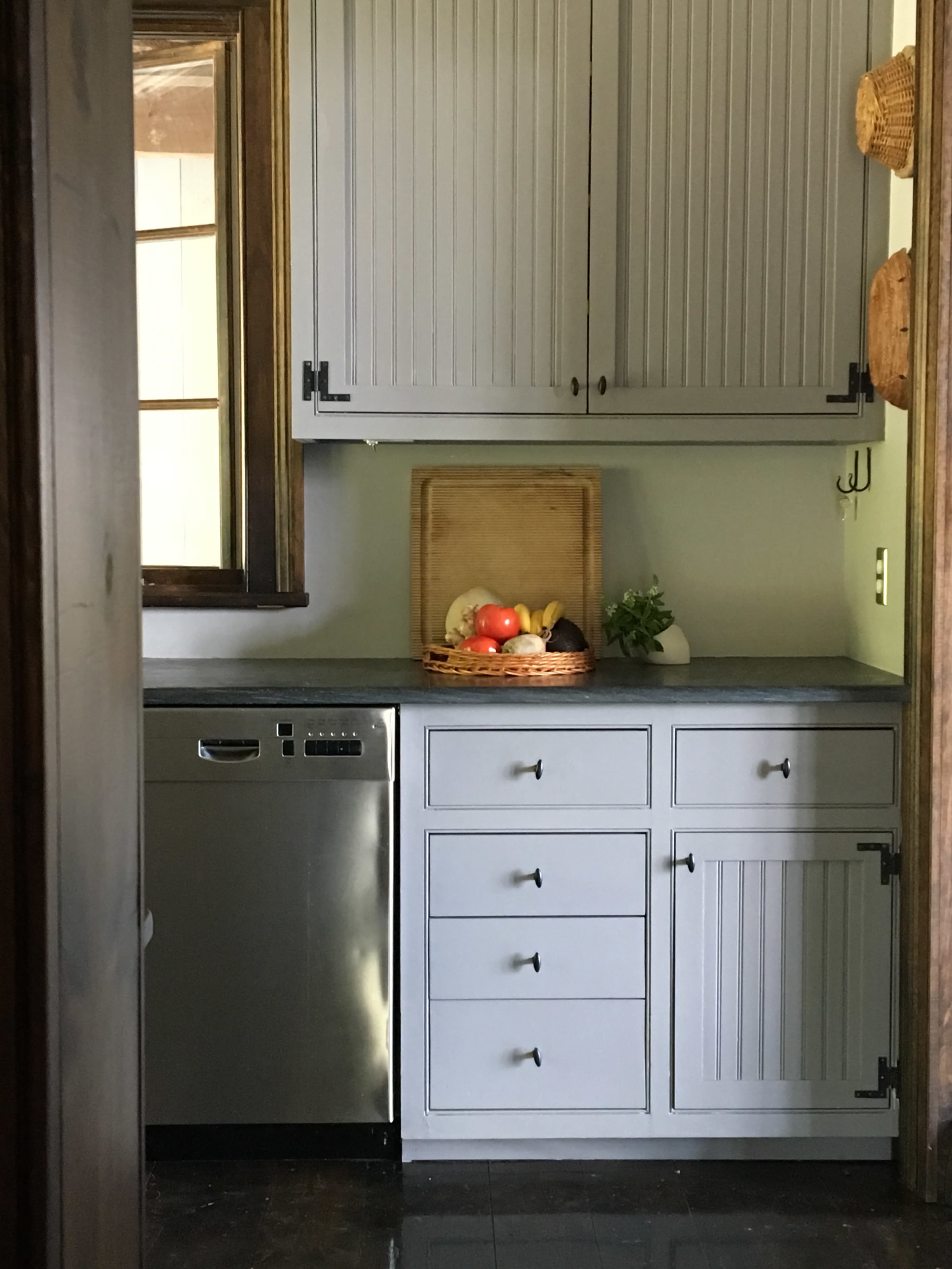 A kitchen features gray cabinets, a stainless steel dishwasher, and a fruit basket on the countertop.