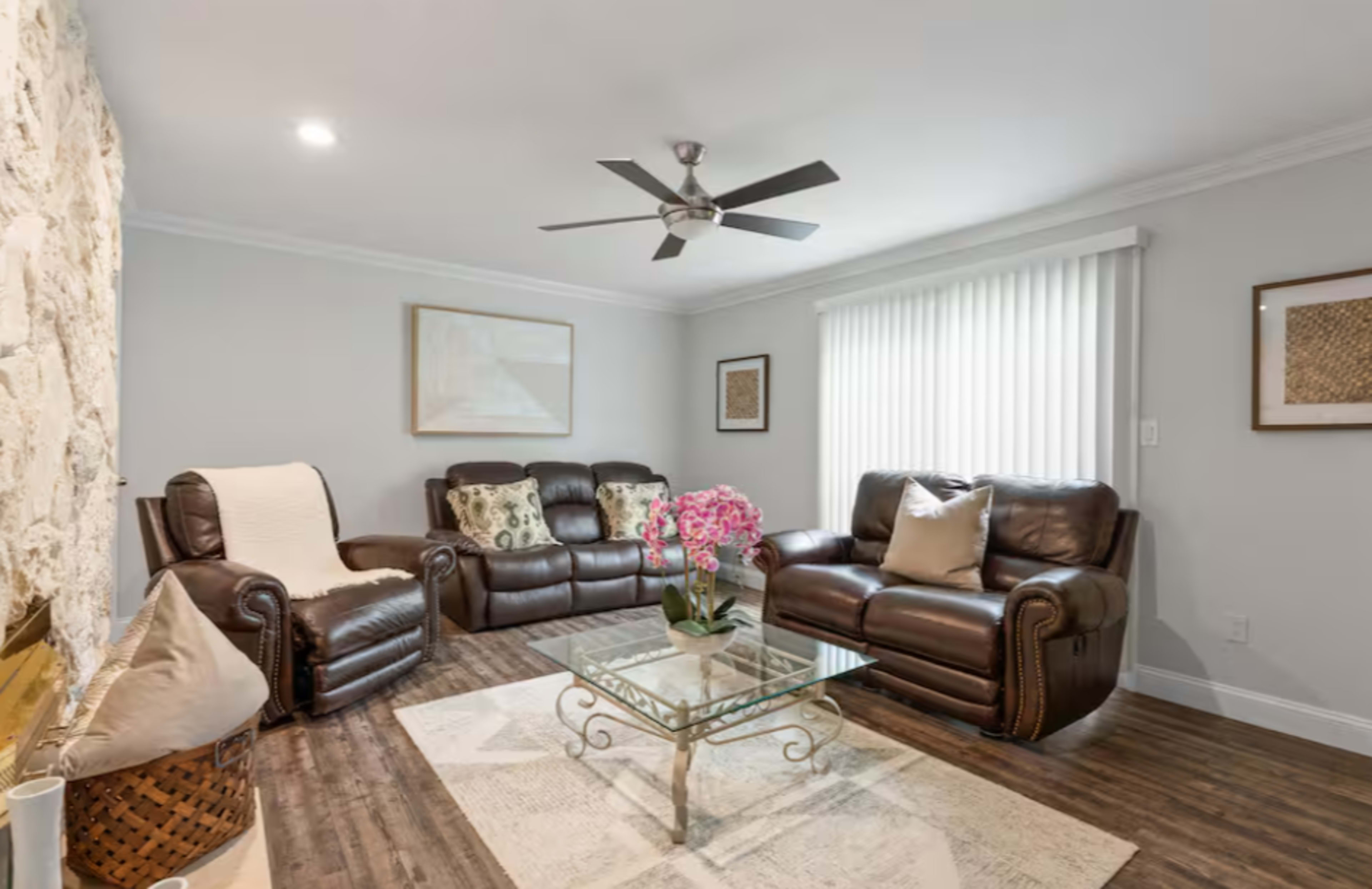 The image shows a living room with two brown leather sofas, a glass coffee table, a textured wall, and a ceiling fan.