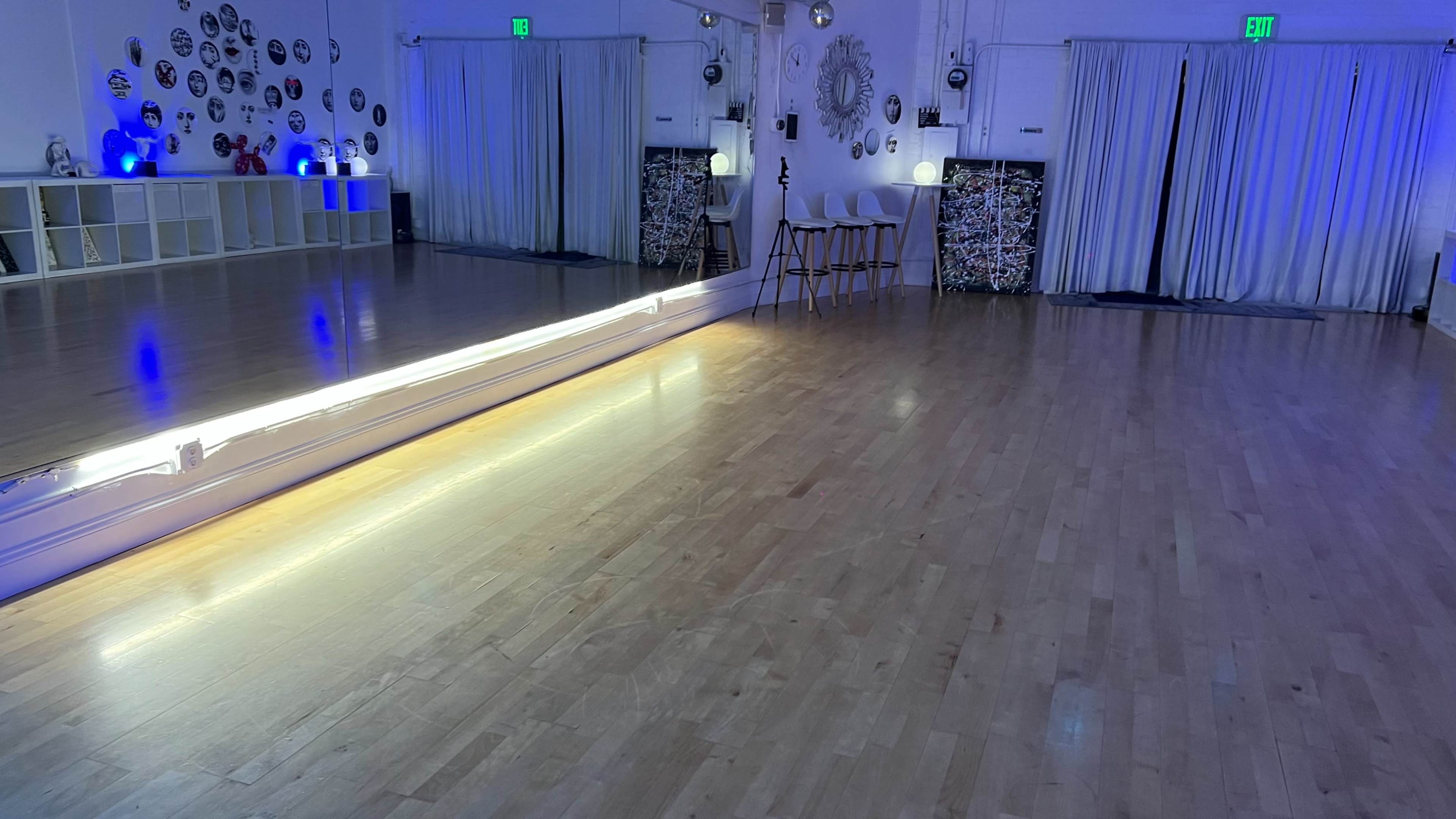 A dance studio with a wooden floor, a large mirror, and blue lighting, along with a seating area featuring three chairs.