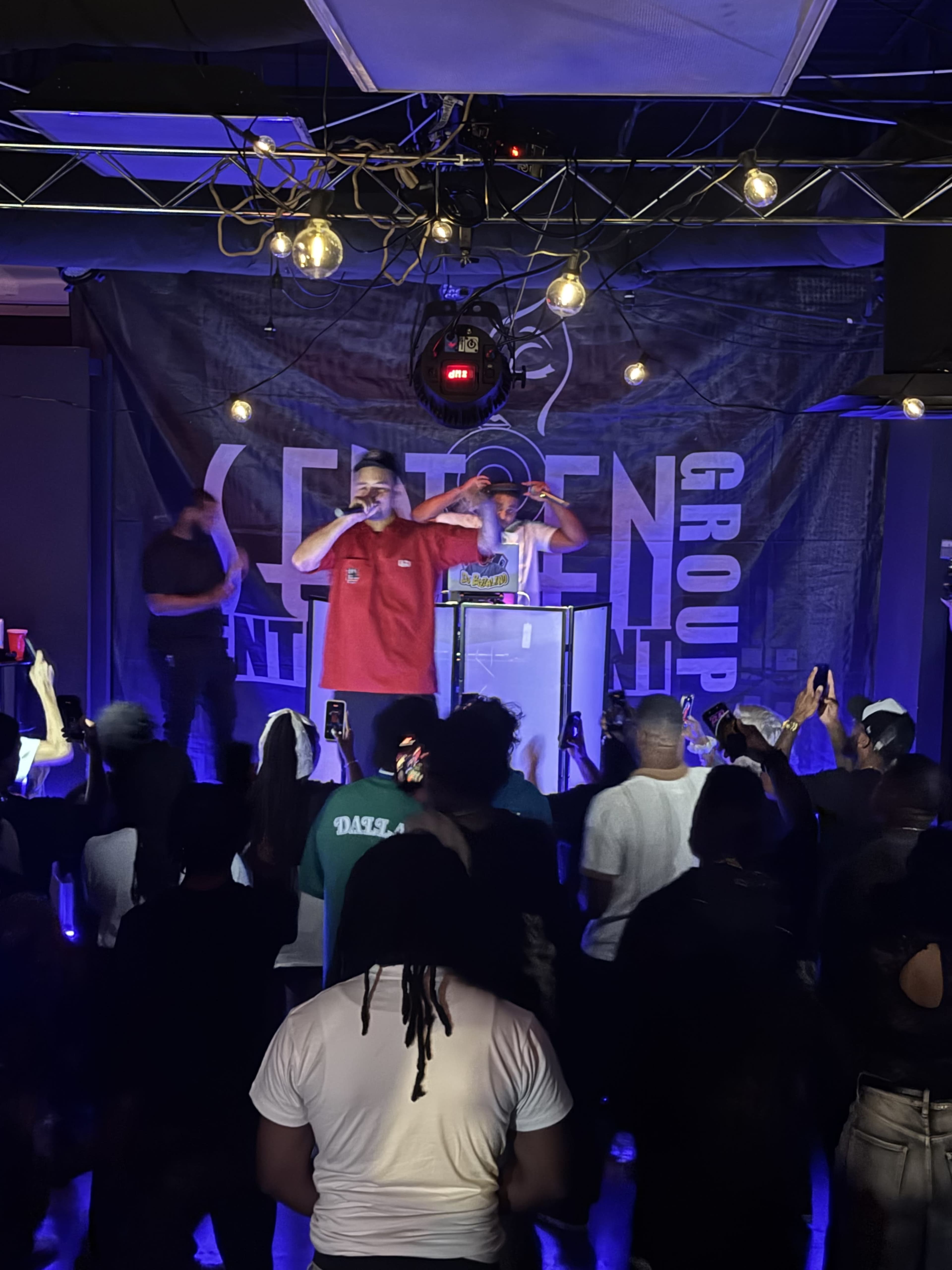 Two performers are on stage in a nightclub, engaging with an audience holding up their phones while the background features a promotional banner for an entertainment group.
